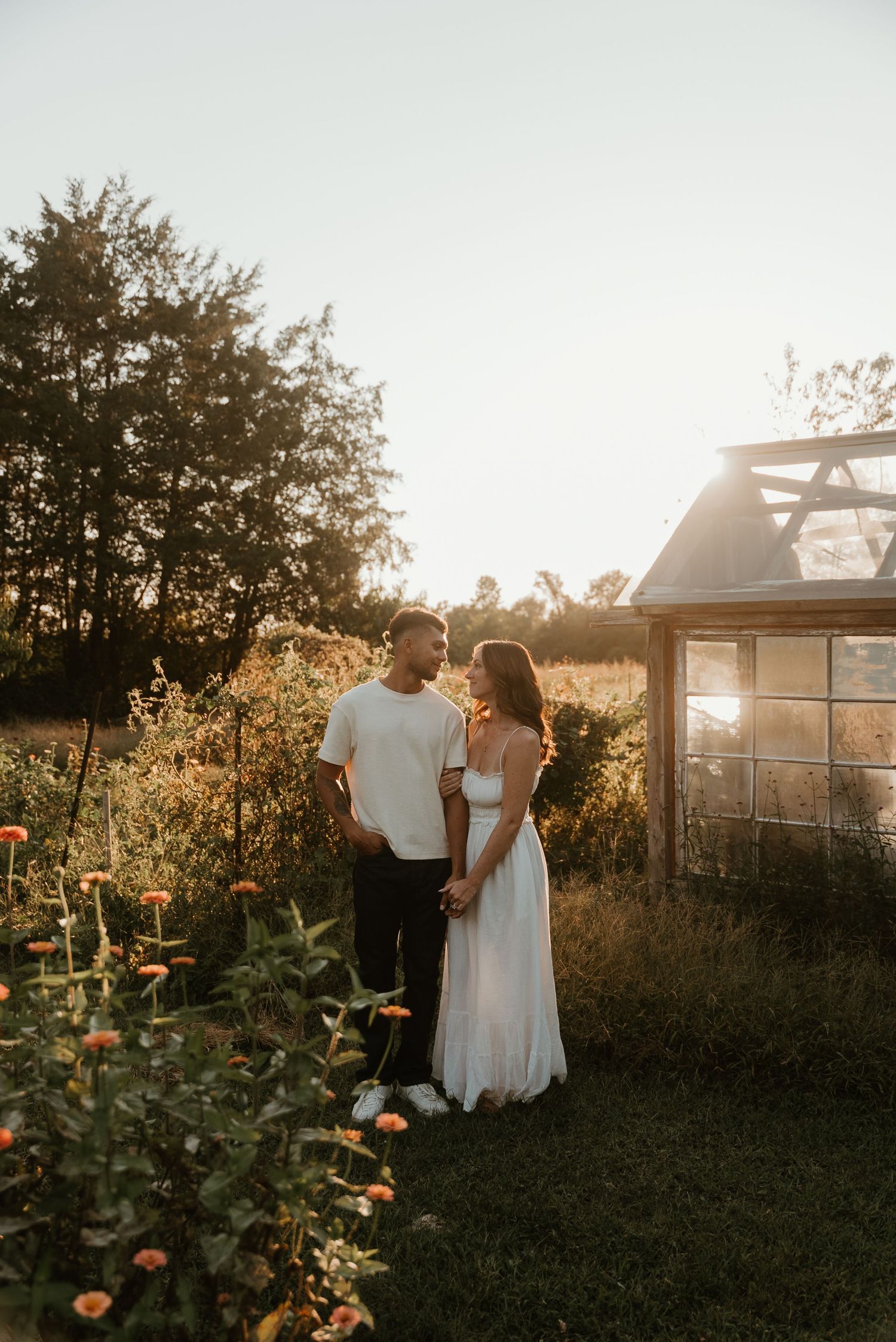 Engaged couple holding hands near a greenhouse surrounded by blooming flowers at sunset.
