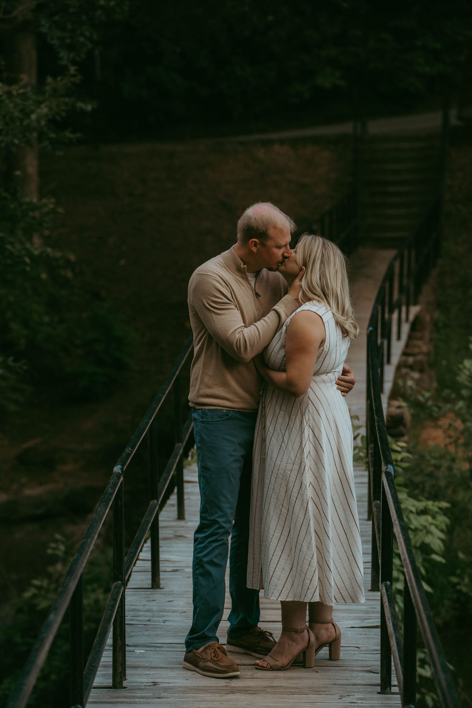 Couple sharing a tender kiss on a wooden bridge surrounded by lush forest greenery.