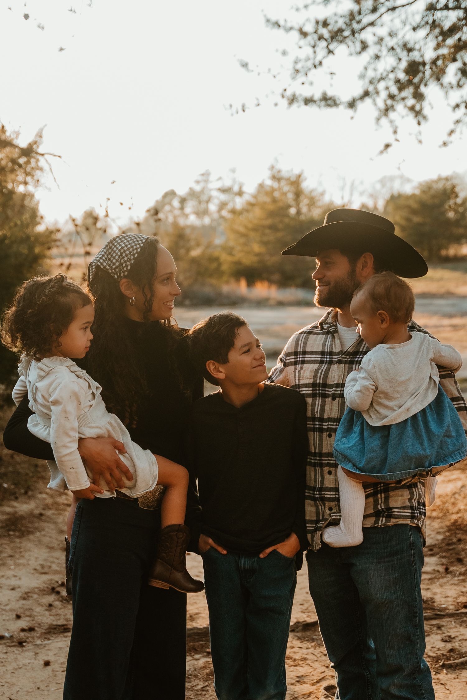 Happy family of five posing outdoors at golden hour, father wearing a cowboy hat holding a baby.