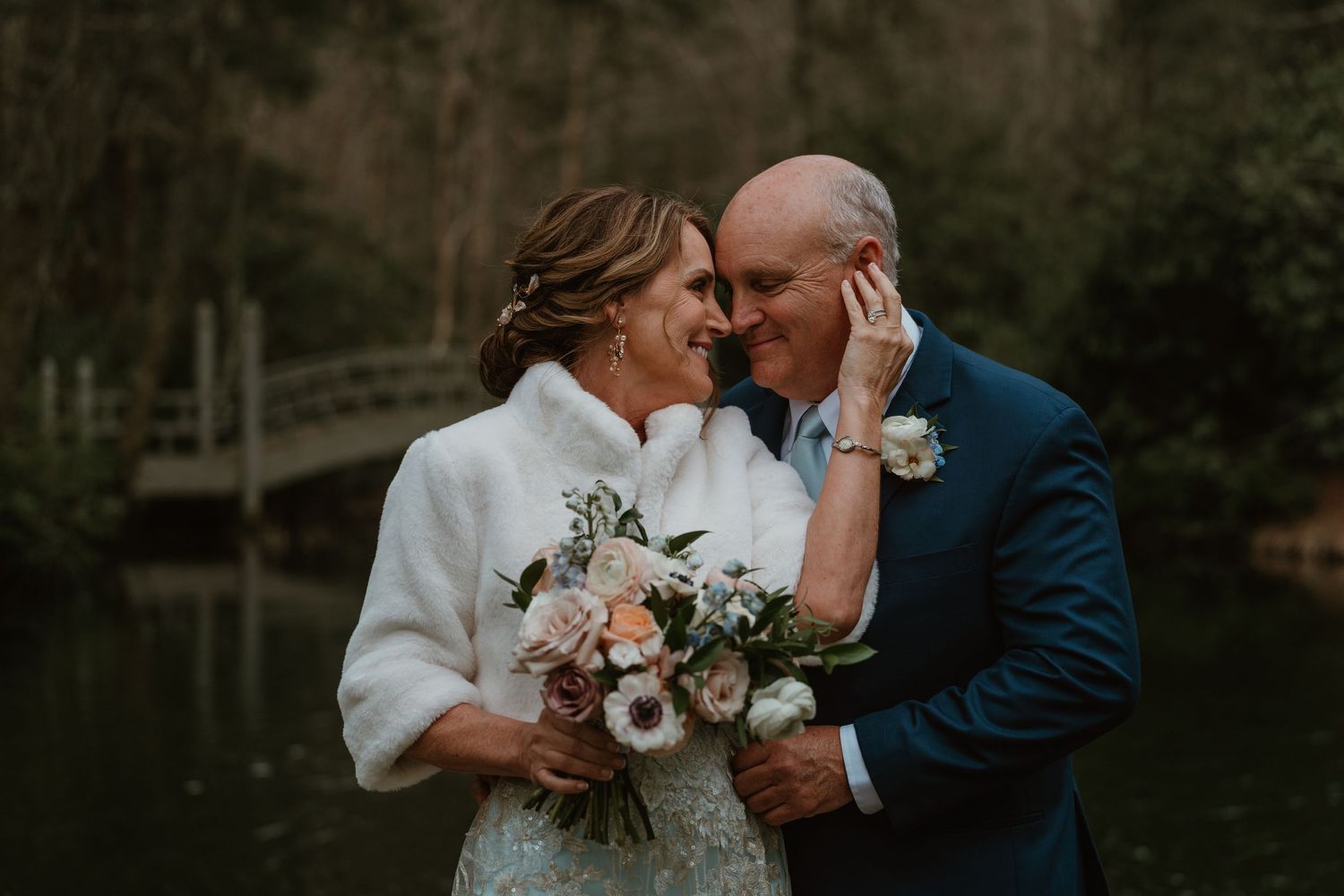 Bride and groom sharing a romantic moment outdoors near a bridge, bride holding a bouquet wearing a white fur wrap.
