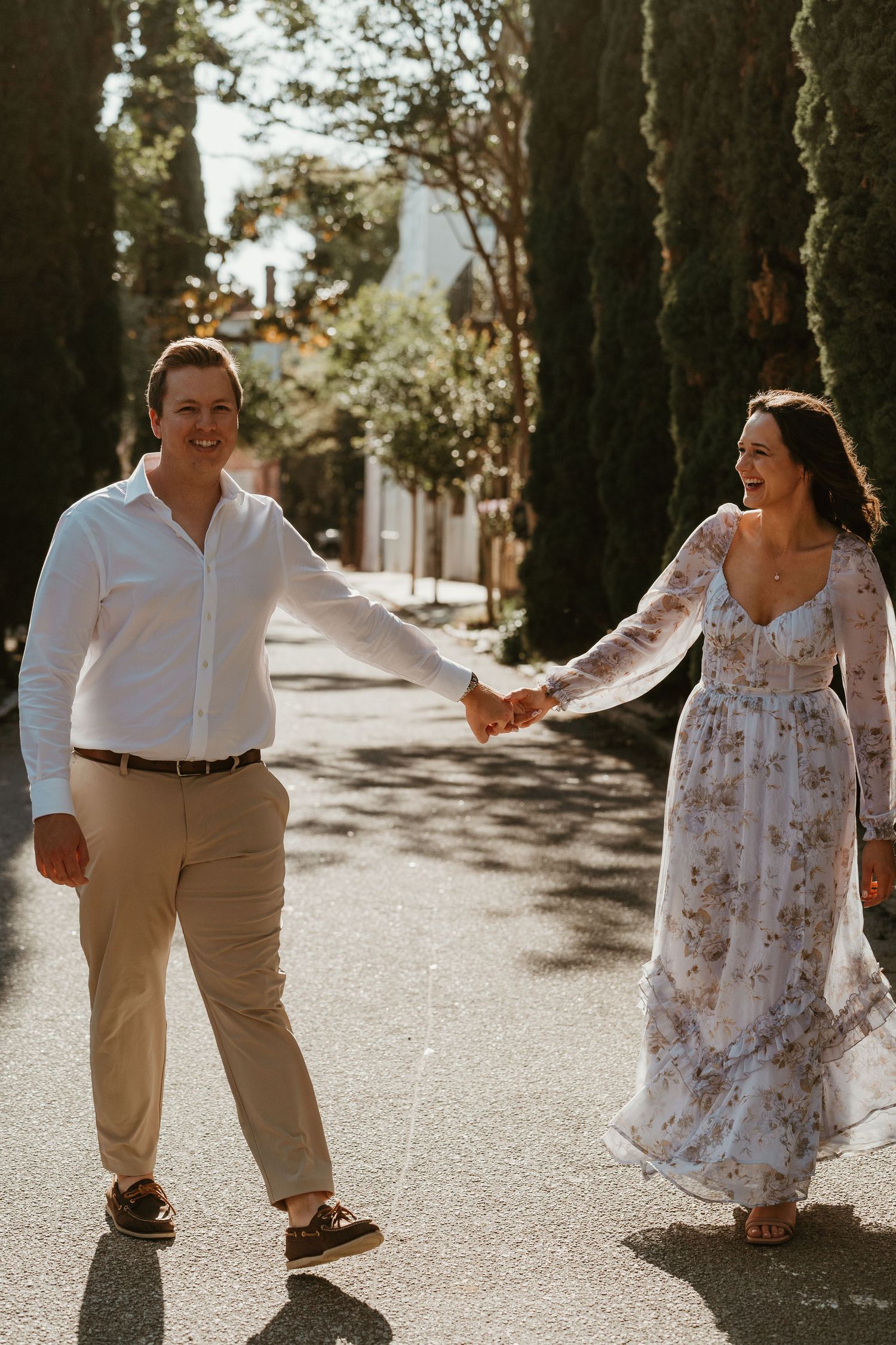 Smiling couple holding hands and walking down a sunny tree-lined street during a golden hour engagement session.