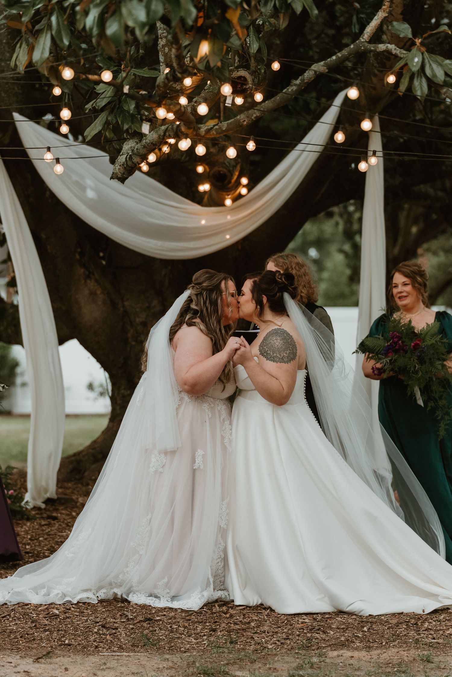 Two brides kiss at altar beneath draped fabric and string lights during outdoor wedding ceremony.