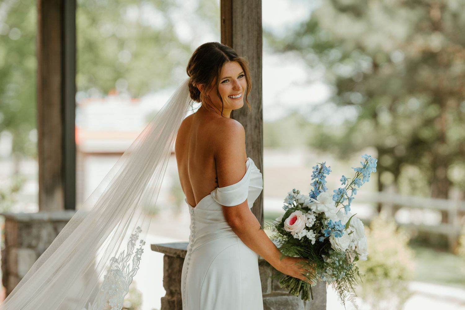 Smiling bride in off-shoulder white gown holds blue and pink bouquet while wearing a flowing veil.