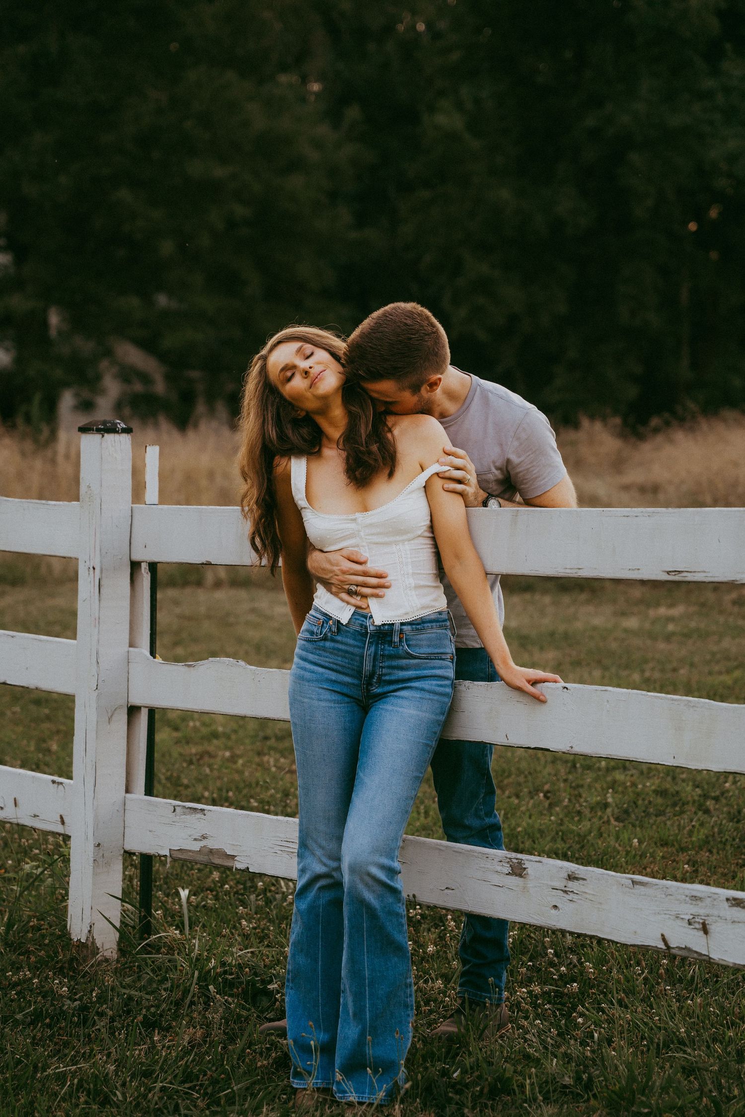 Romantic couple embracing by a white wooden fence in a countryside field at golden hour.