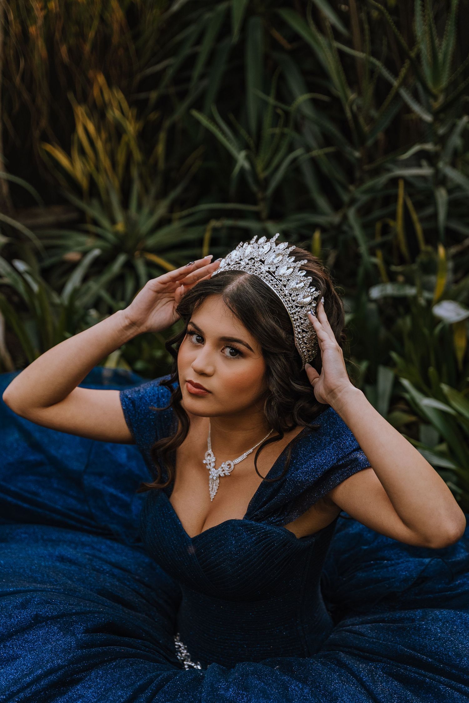 A young woman in a sparkling navy ball gown adjusts her crystal crown while seated among green plants.