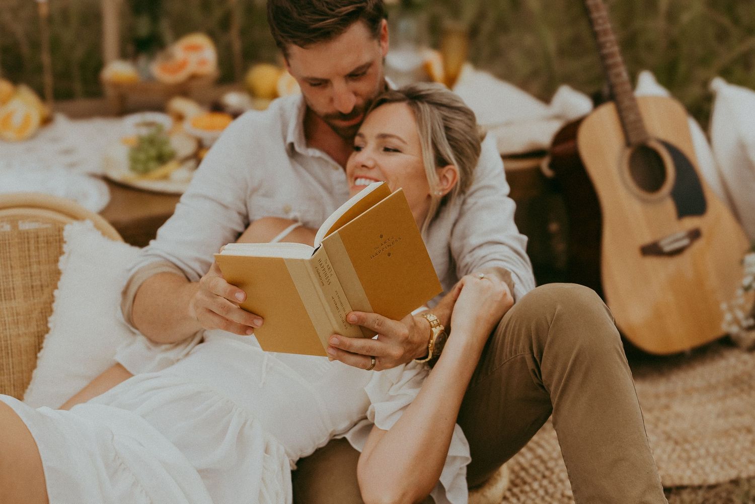 Couple cuddling and reading a yellow book together at a romantic outdoor picnic with a guitar nearby.