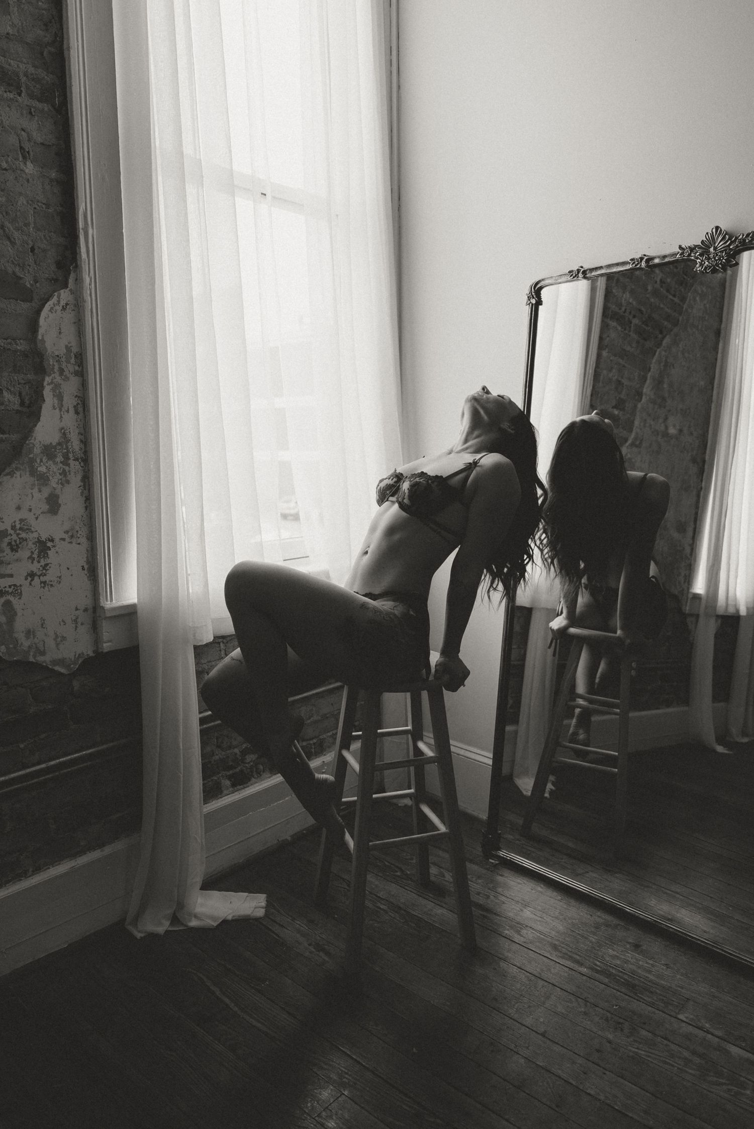 Black and white boudoir photo of woman sitting on stool by bright window with mirror reflection.
