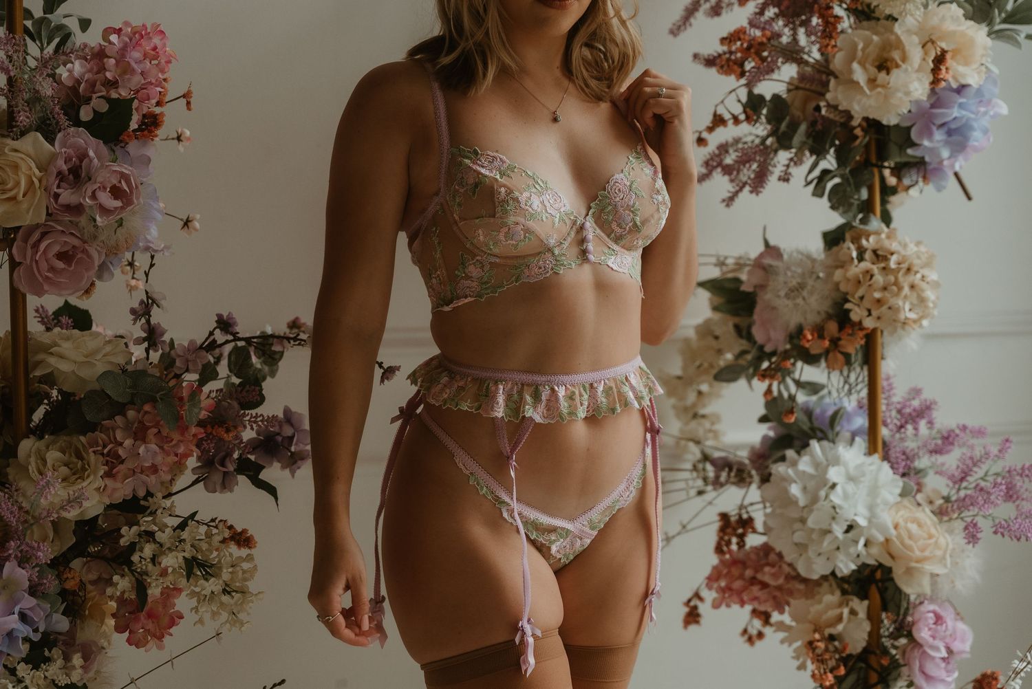 Woman wearing sheer floral embroidered lingerie set with garter belt surrounded by flowers.