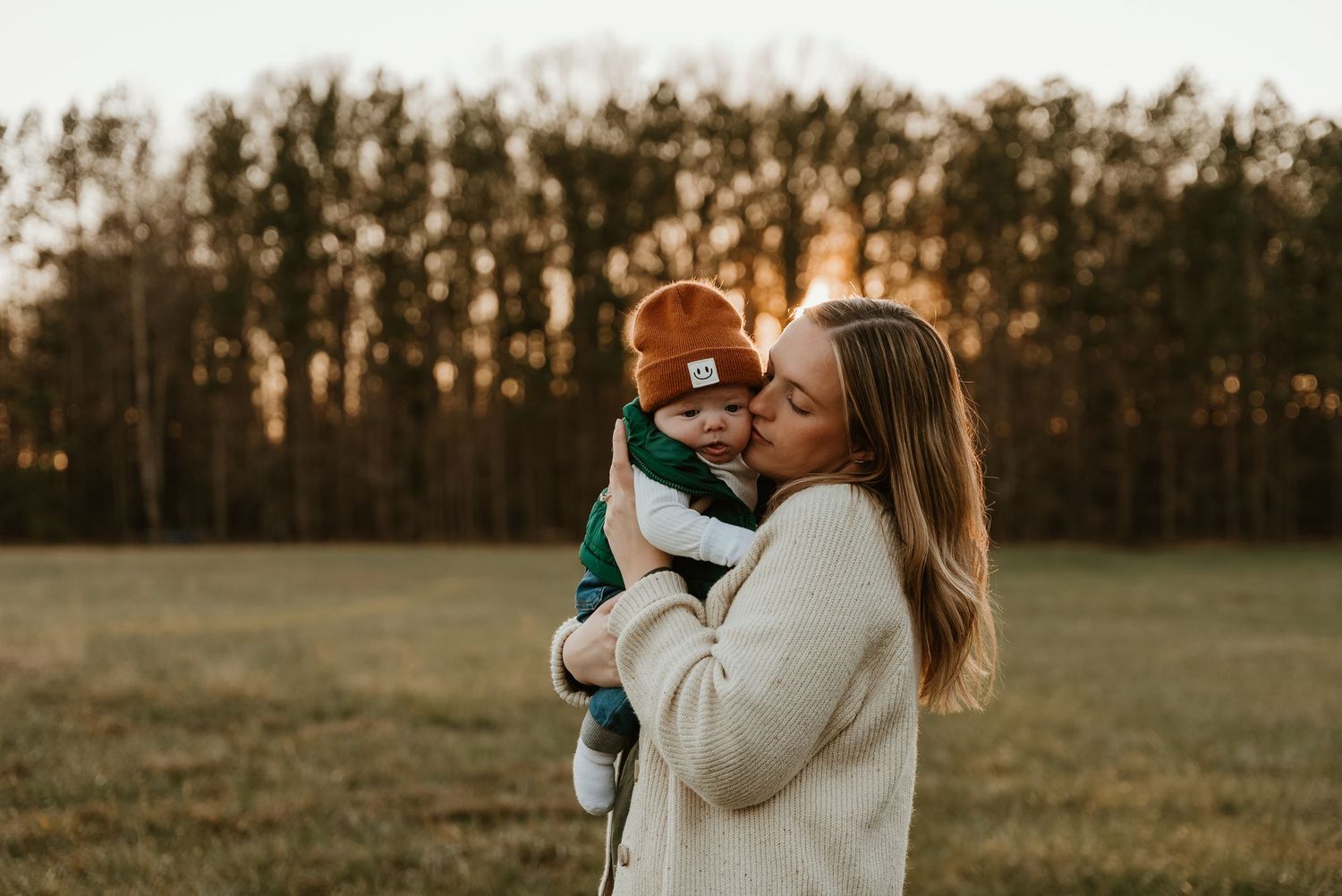 A mother tenderly kisses her baby wearing a rust beanie during a golden hour outdoor photo session.