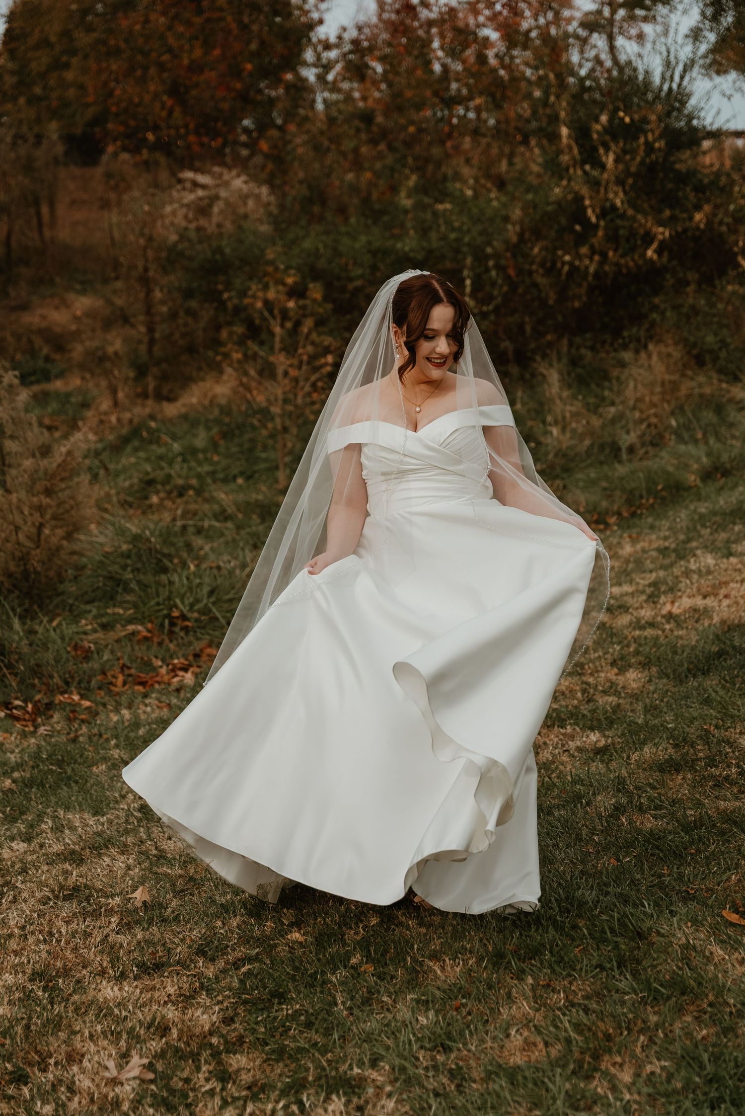 Bride in an off-shoulder white gown and veil twirling in an autumn outdoor setting.