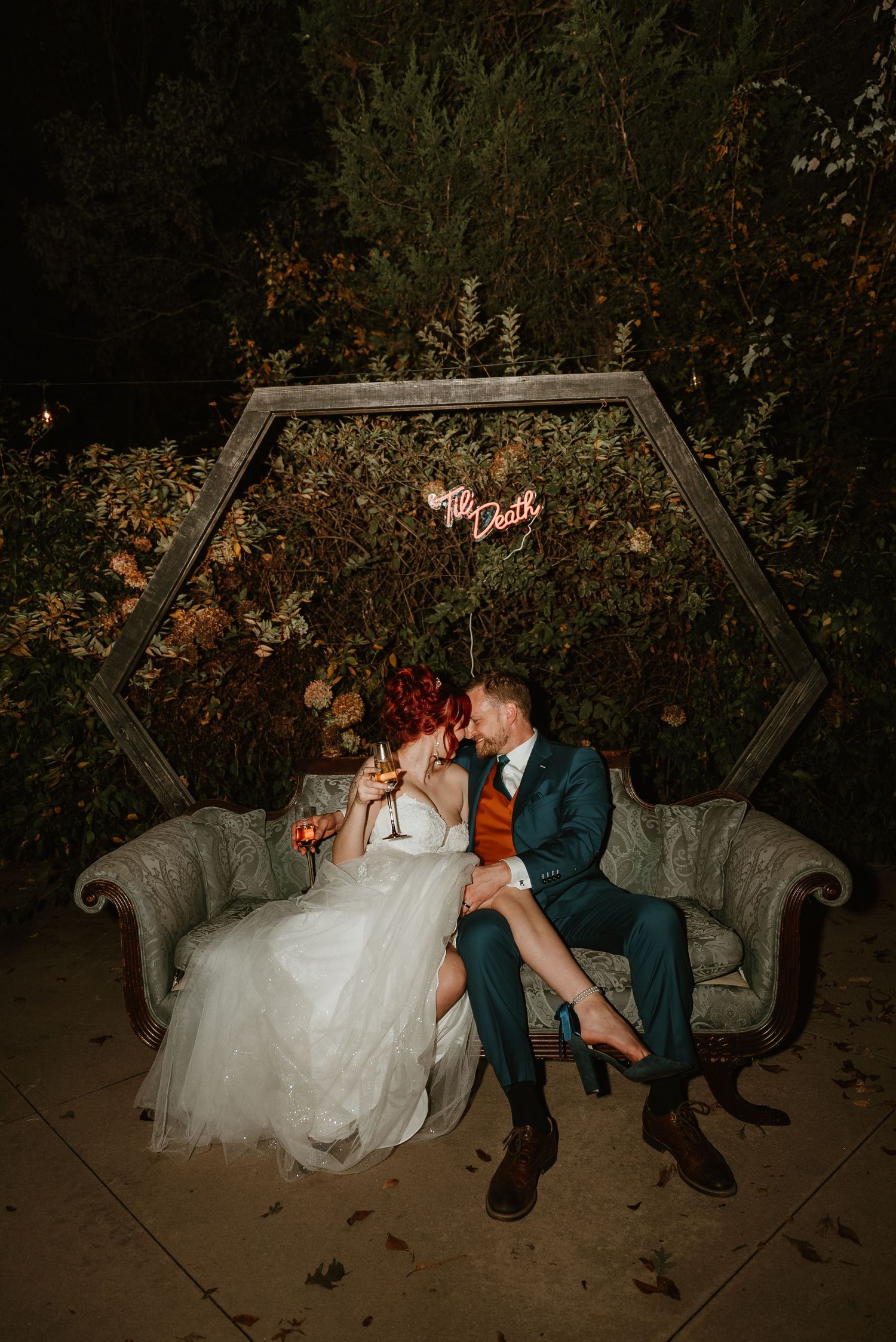 Wedding couple sits on vintage sofa under hexagonal arch with neon sign at evening reception.