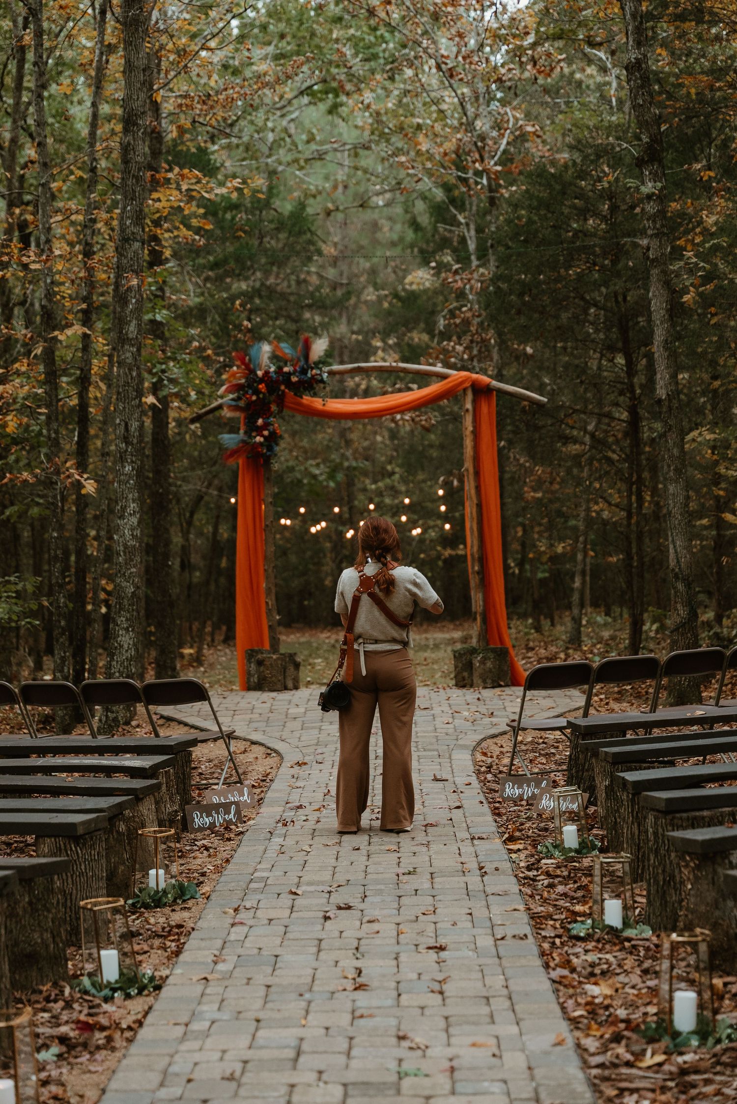Wedding photographer walking down brick aisle toward rustic fall arch with orange draping and string lights.