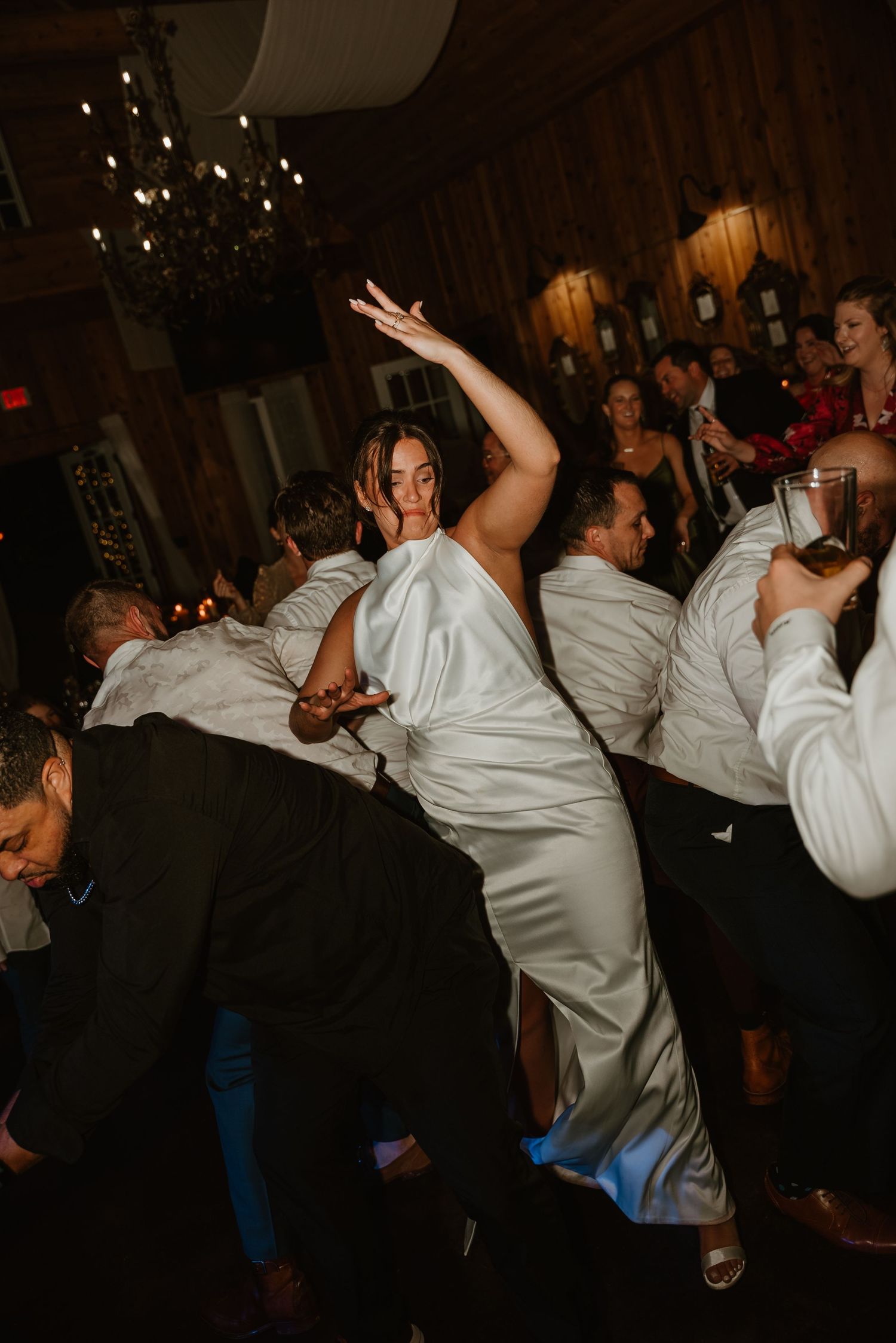 Bride in sleek white gown dances energetically with wedding guests on a dimly lit reception dance floor.