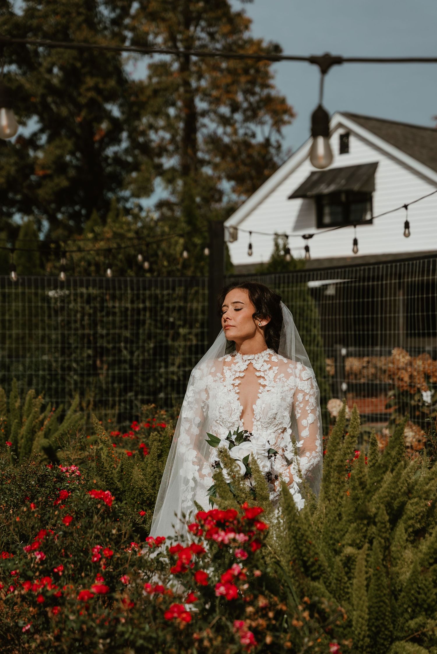 Bride in lace long-sleeve wedding gown with veil standing among red flowers and greenery in garden.