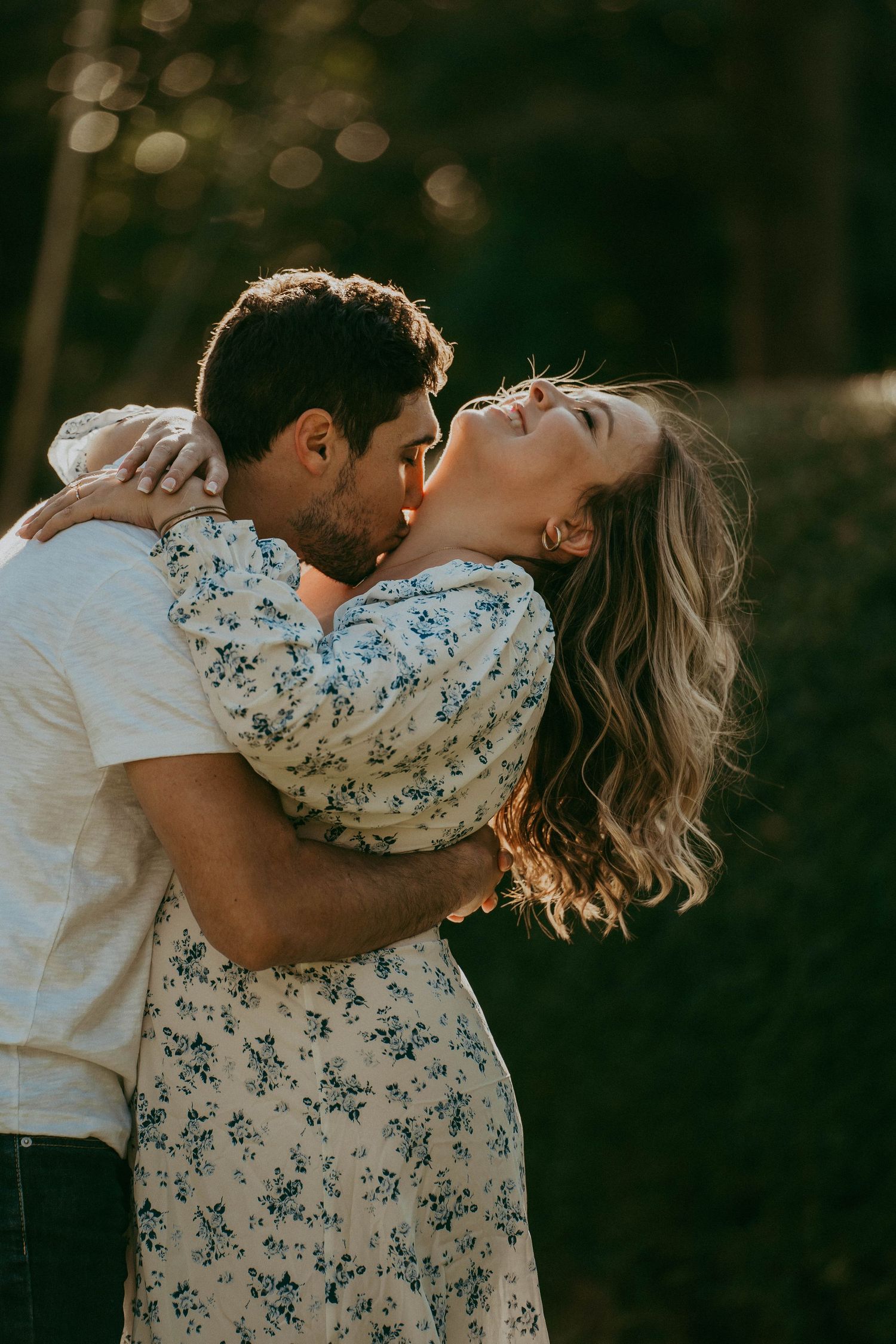 Romantic couple kissing outdoors at golden hour, woman in floral dress leaning back joyfully.