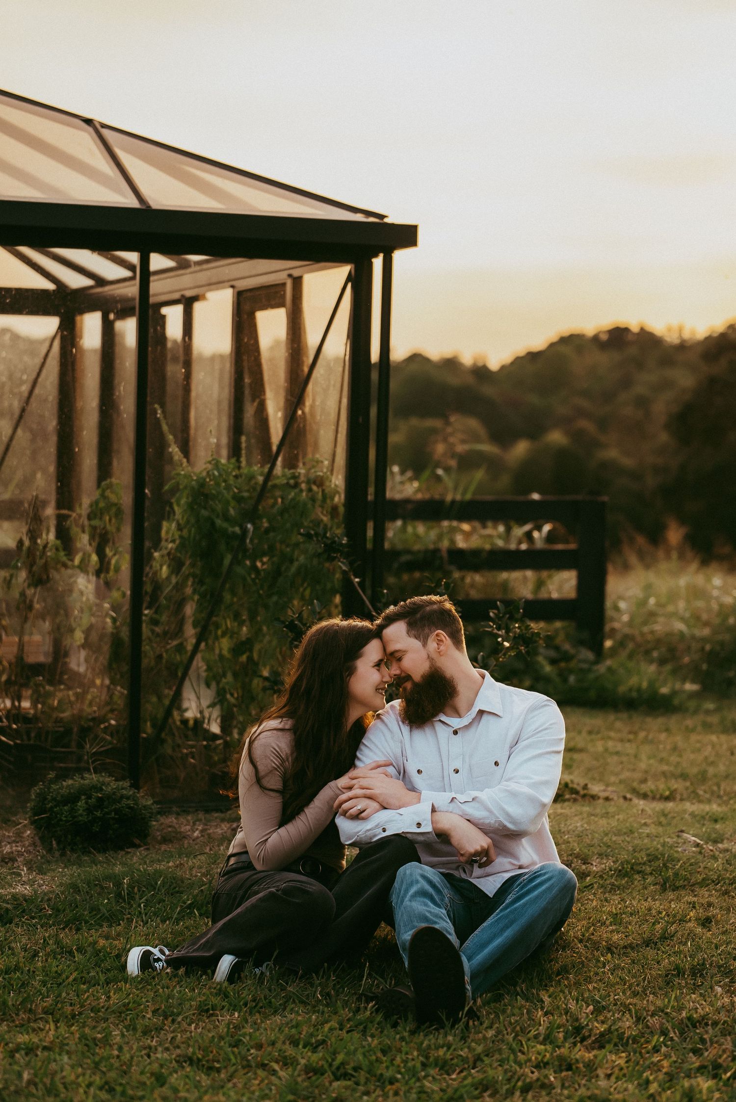 Couple sitting close on grass near greenhouse at sunset, leaning in for a kiss.