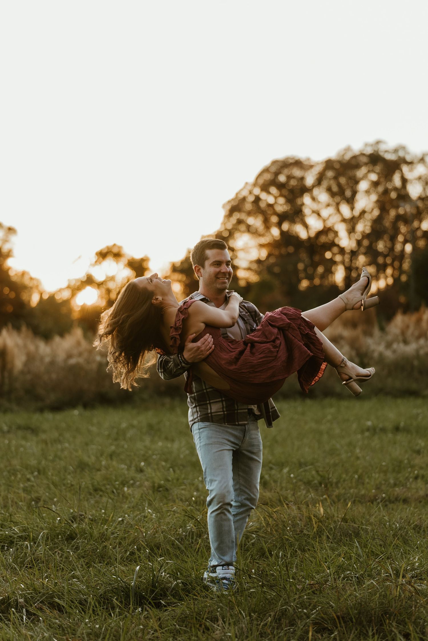 Man lifting woman in his arms in a golden sunset field, both laughing joyfully during engagement shoot.