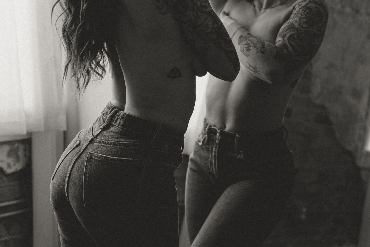 Black and white artistic boudoir photo of two tattooed women in jeans posing near window.