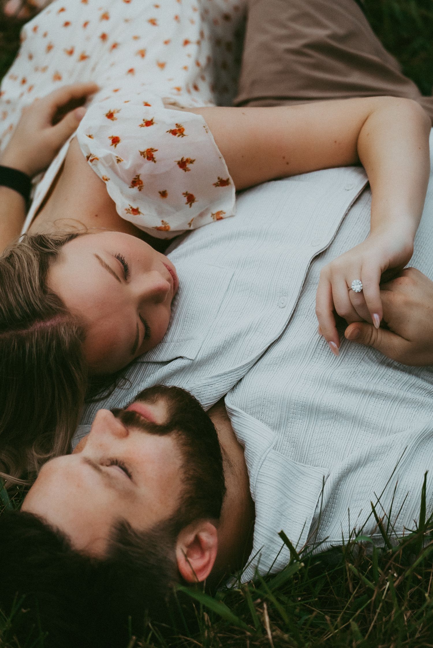 Couple lying in grass with eyes closed, woman resting head on man's chest showing engagement ring.