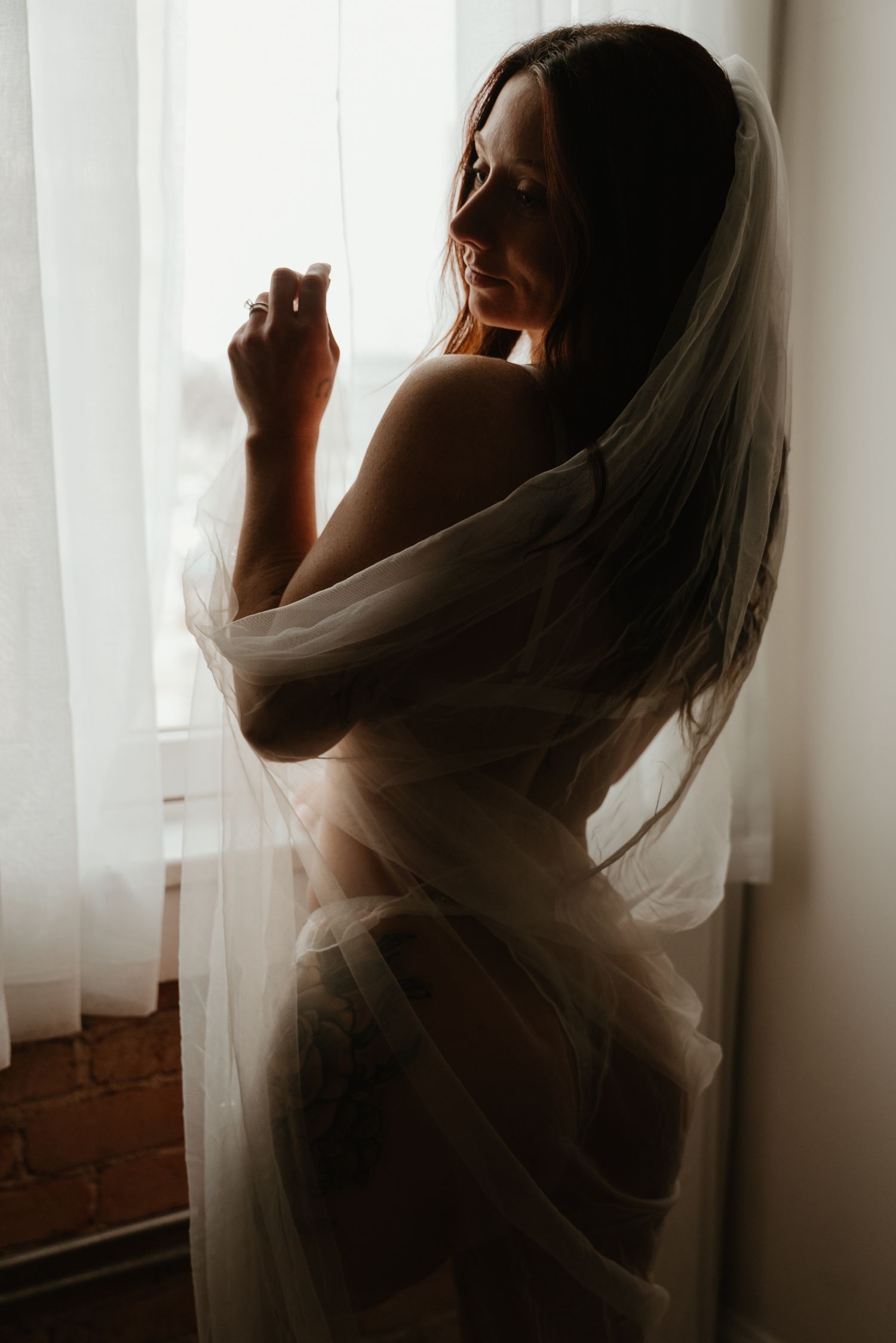 Silhouette of woman wrapped in sheer white tulle fabric standing by bright window in artistic boudoir photo.