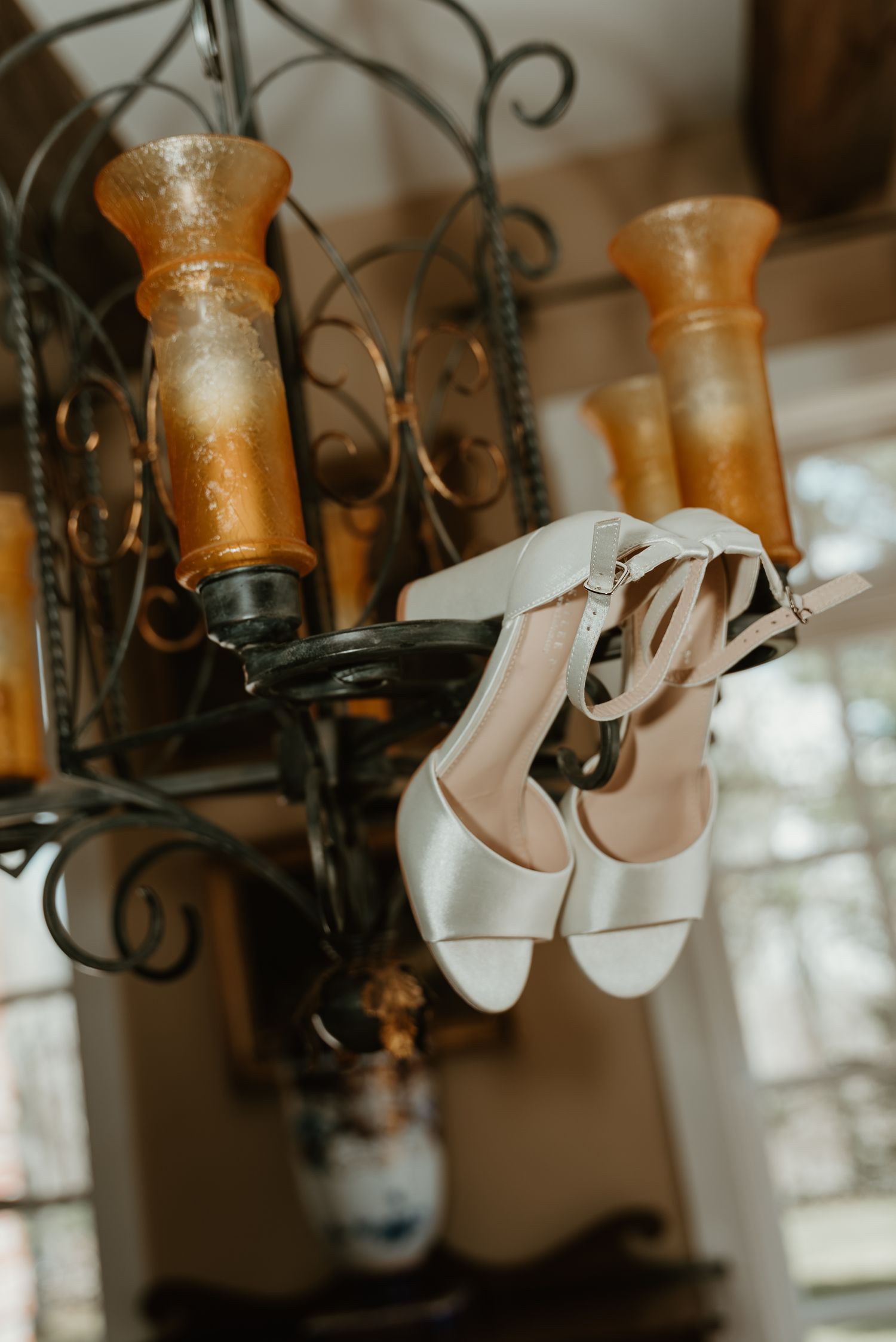 White bridal heels with ankle straps hang from a rustic iron chandelier with amber glass candle holders.