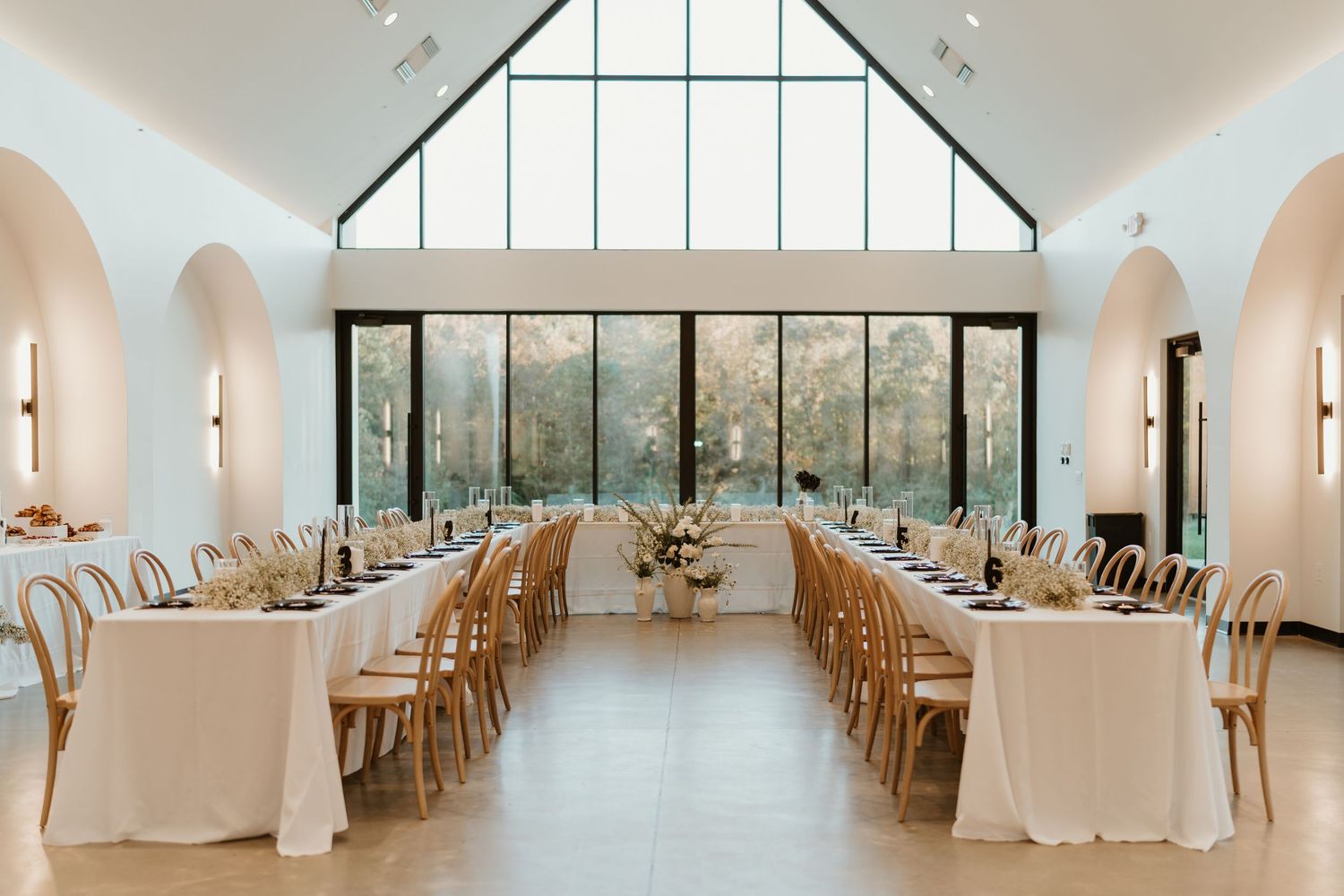 Elegant minimalist wedding reception hall with long white tables, wooden chairs, and floor-to-ceiling windows.