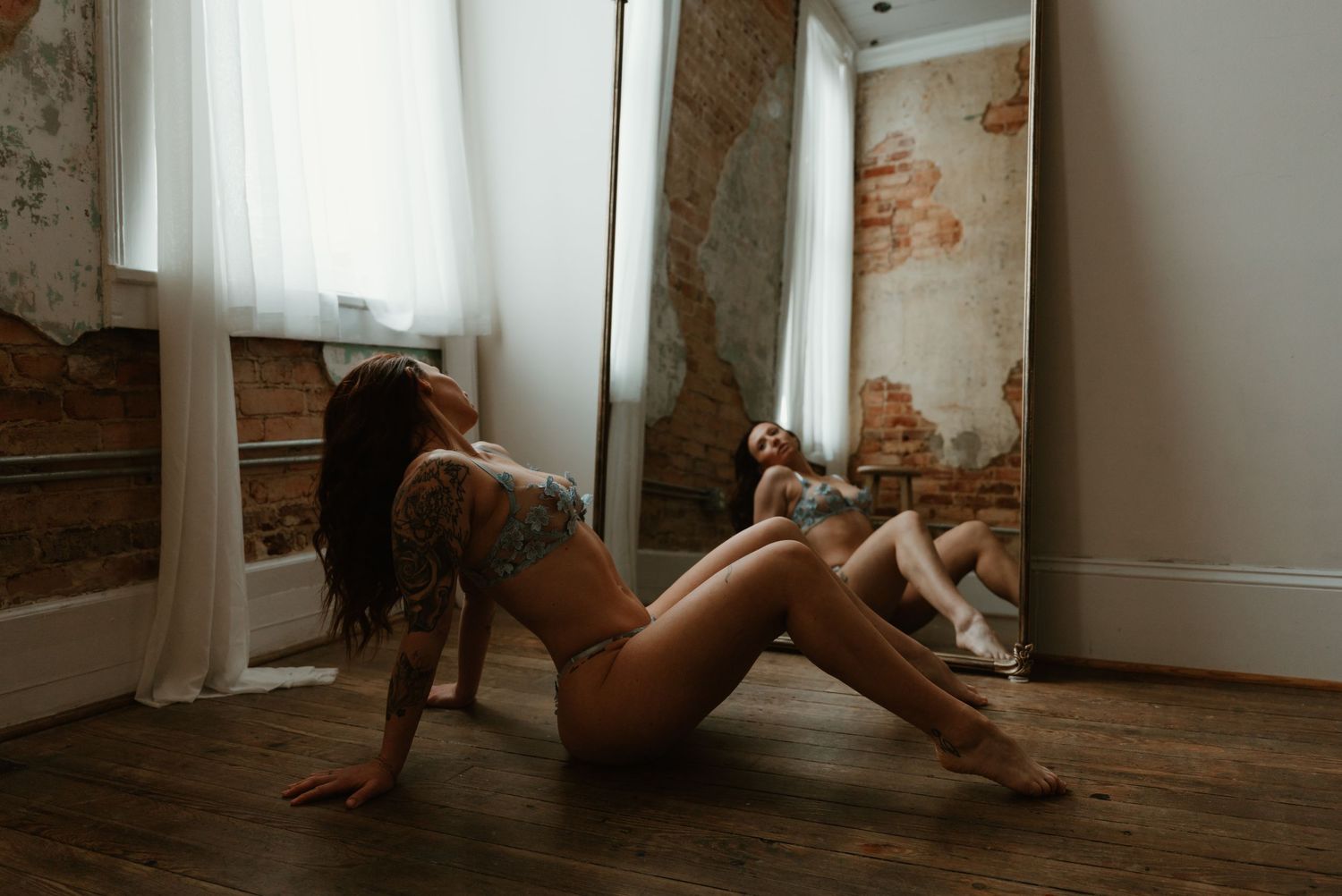 Tattooed woman in nude lingerie sits on hardwood floor beside a large mirror in rustic studio.