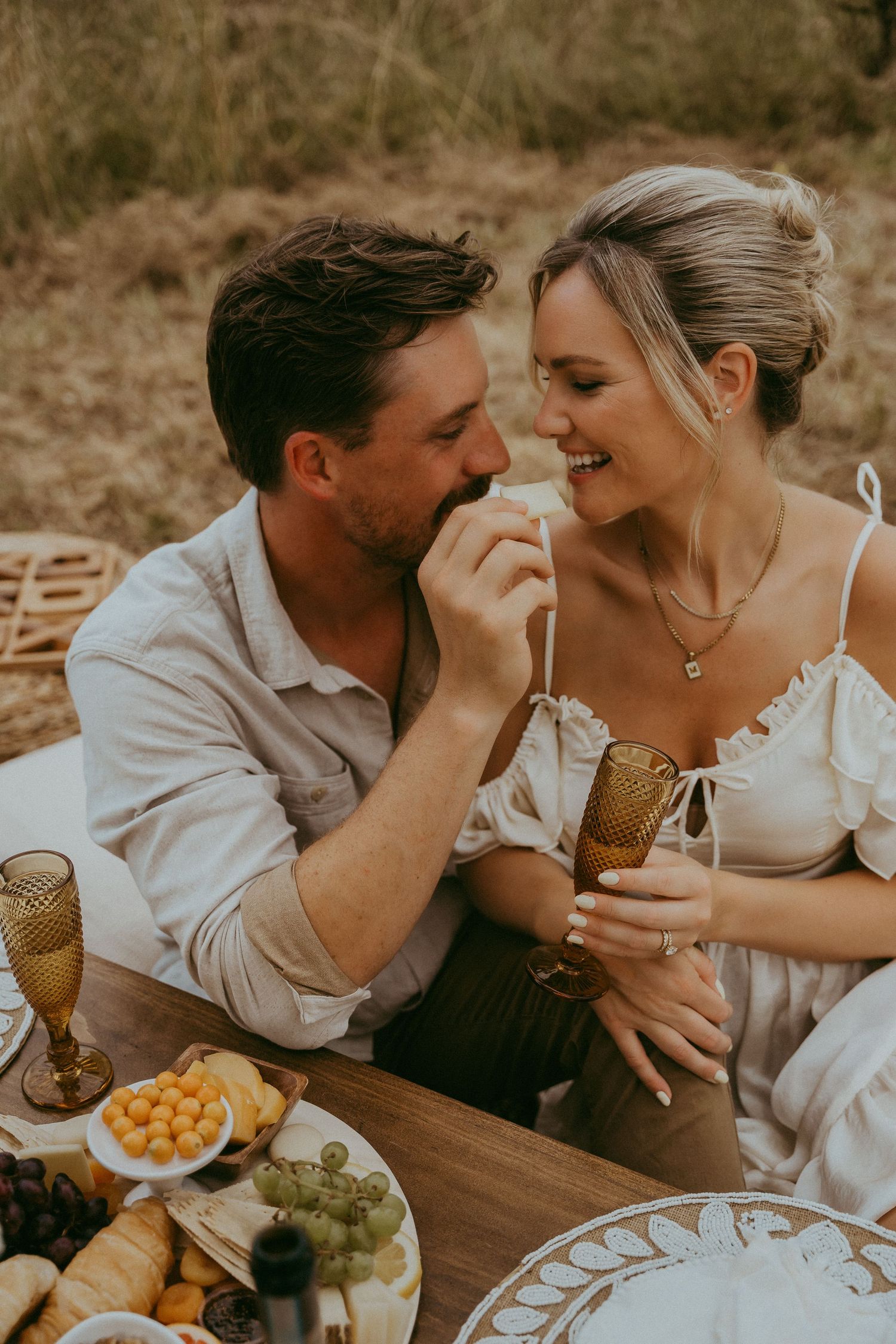 Happy couple sharing food at a romantic outdoor picnic with grapes, cheese, and amber glassware.