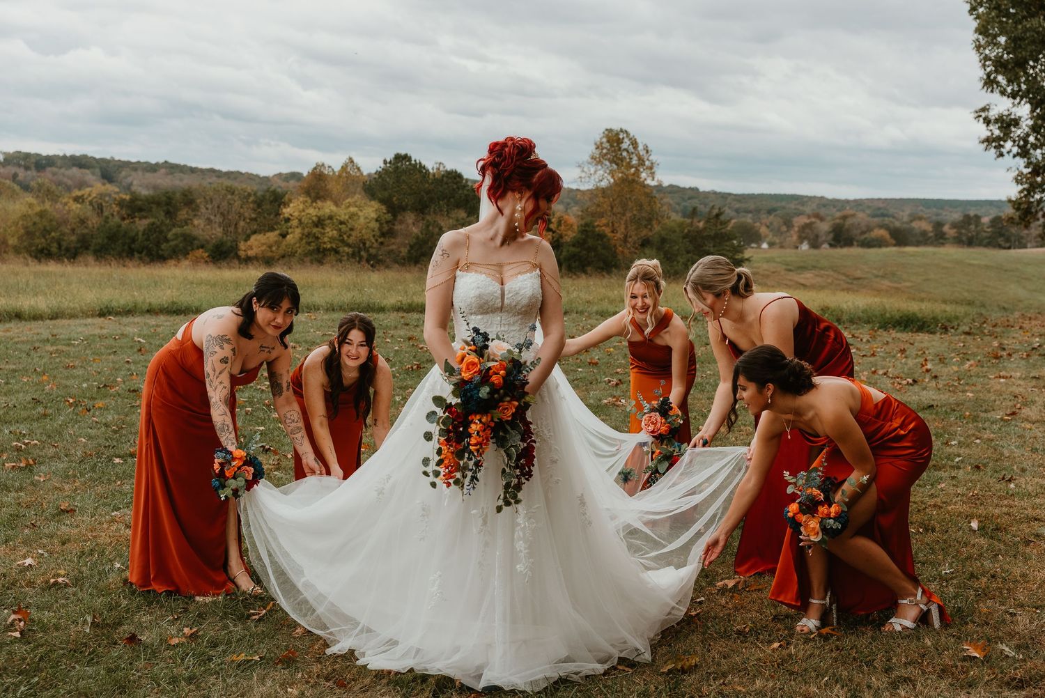 Bridesmaids in rust orange dresses adjust bride's white gown in scenic outdoor fall wedding setting.