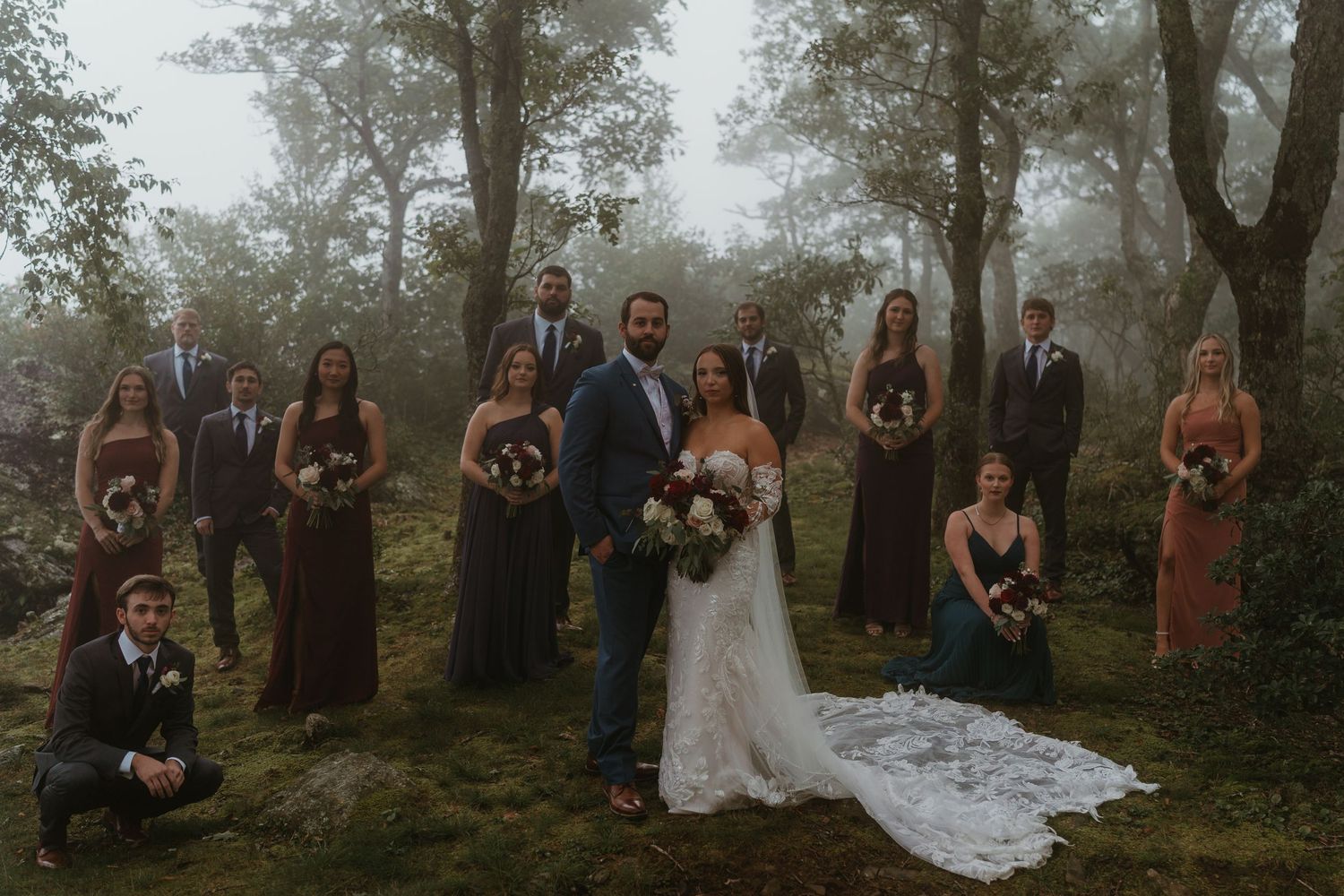 Wedding party posing together in misty forest with bridesmaids in burgundy dresses and groomsmen in navy suits.
