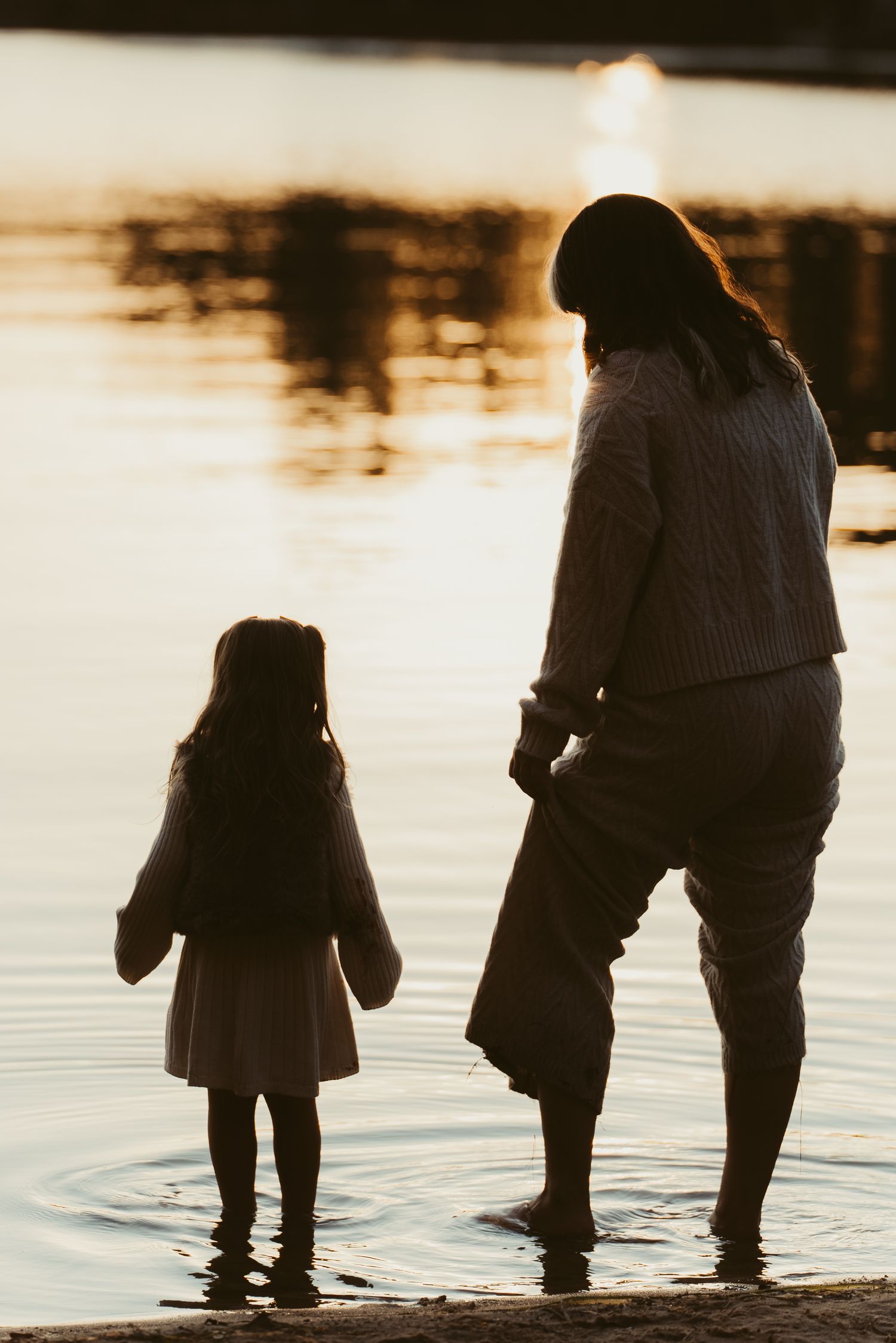 Silhouette of a woman and young girl standing in shallow water at sunset by a tranquil lake.
