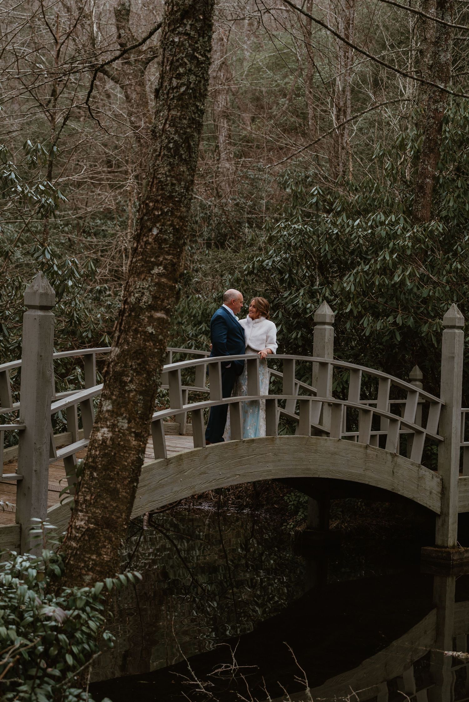 Wedding couple standing together on a wooden arched bridge surrounded by lush forest trees.