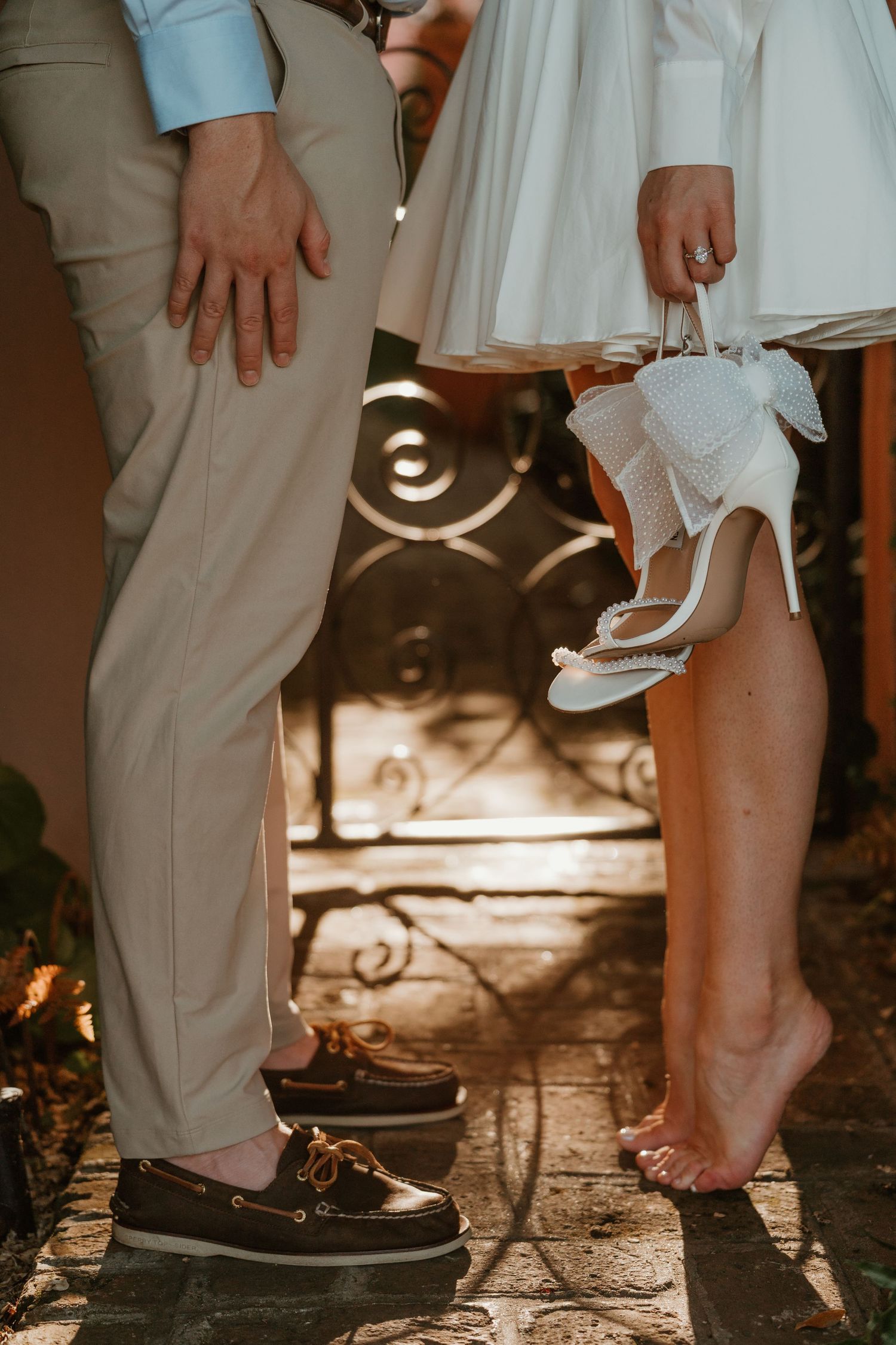 Close-up of couple's feet, man in brown boat shoes and woman barefoot holding white heels on cobblestone.