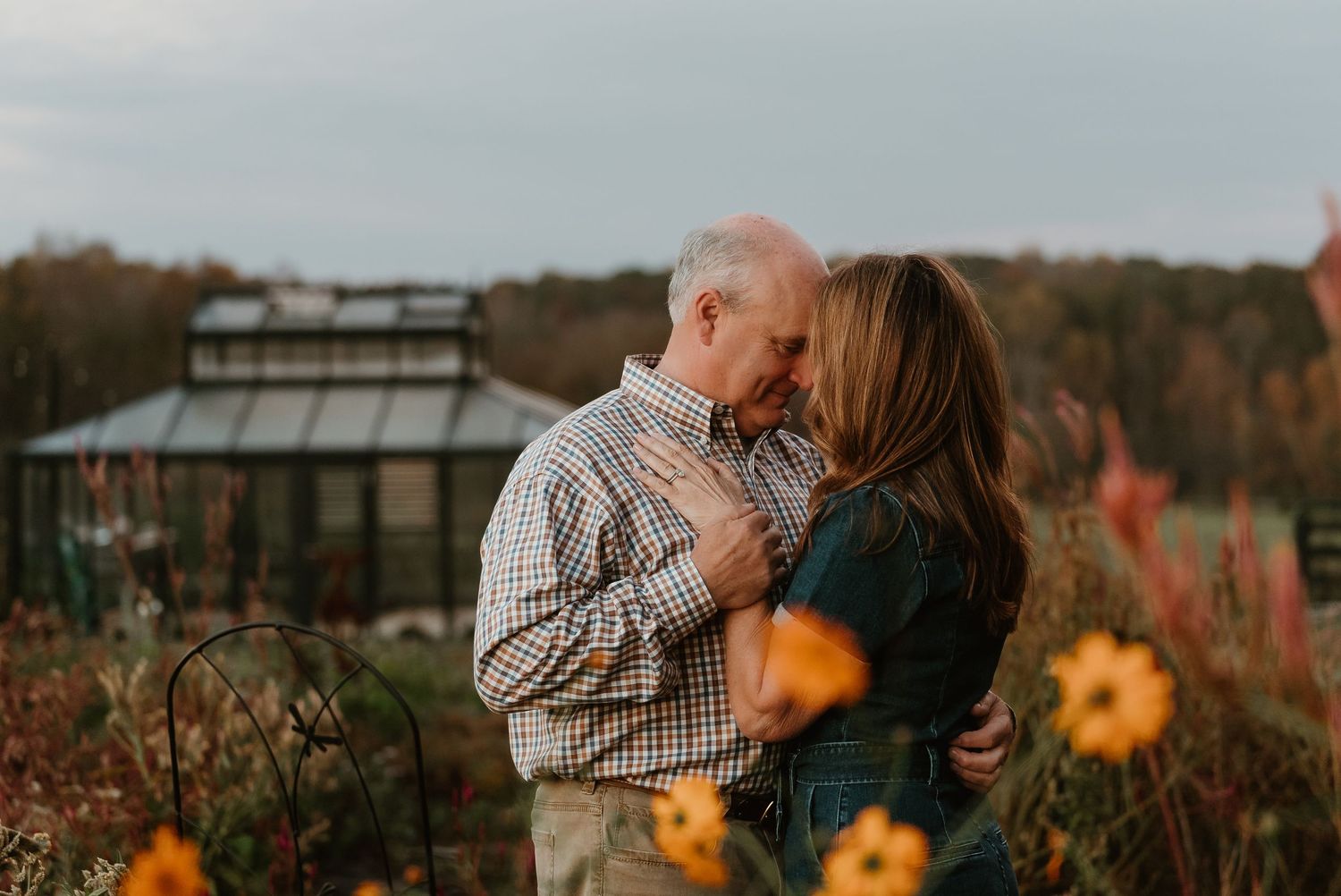 Romantic couple nearly kissing in a garden with autumn foliage and greenhouse backdrop.