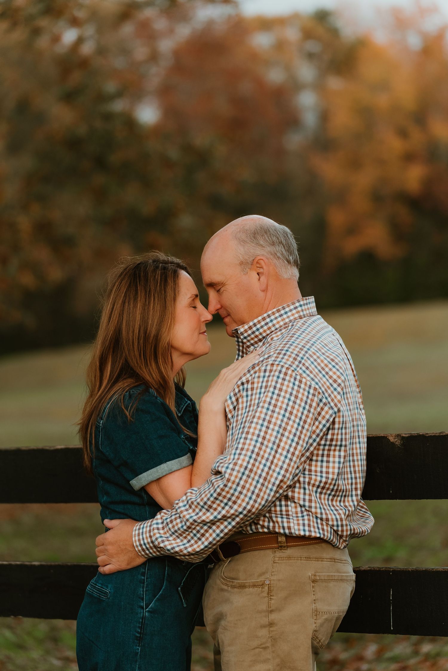 Couple sharing an intimate moment leaning against a wooden fence with autumn trees behind.