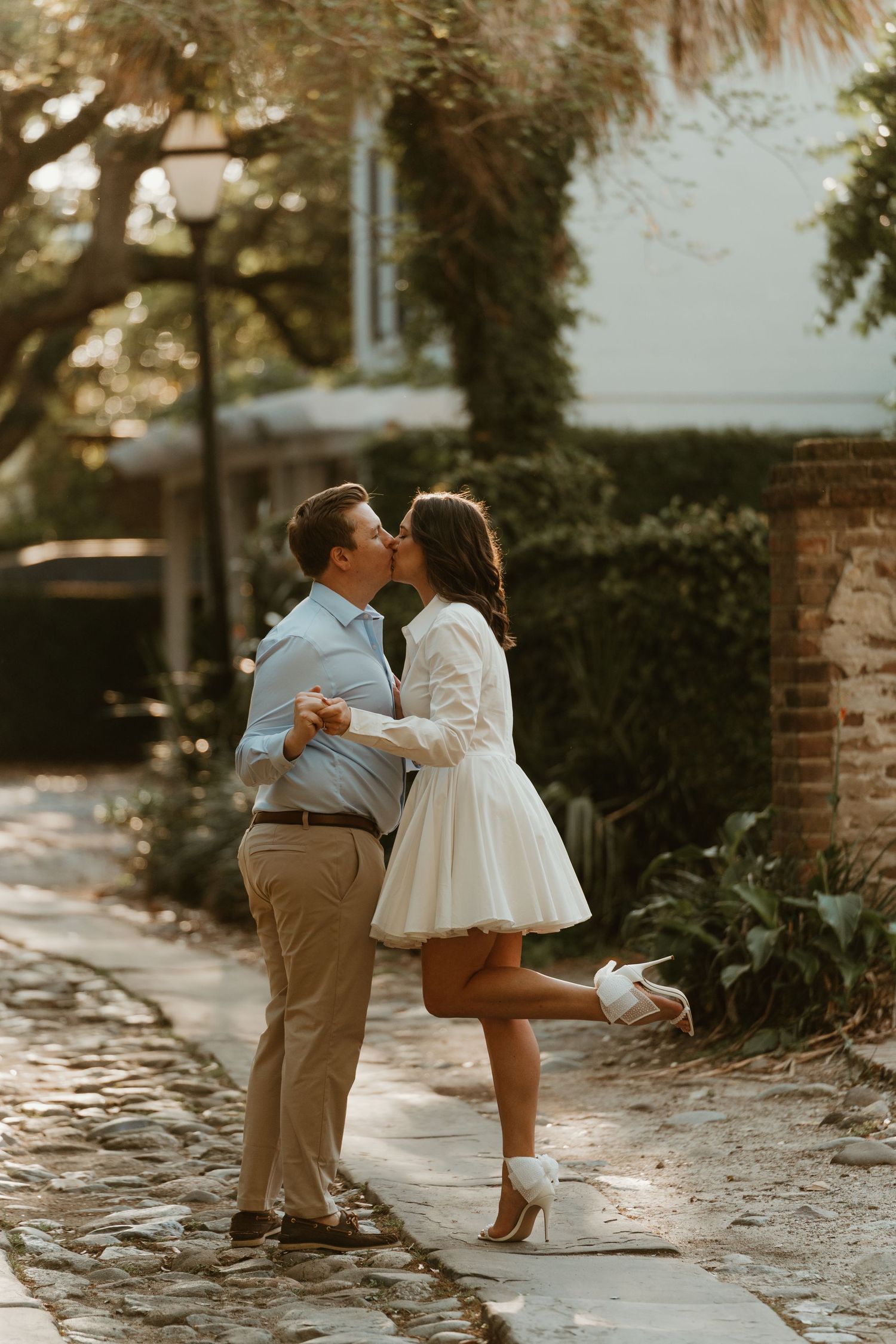 Couple sharing a romantic kiss on a cobblestone path surrounded by lush greenery during golden hour portraits.