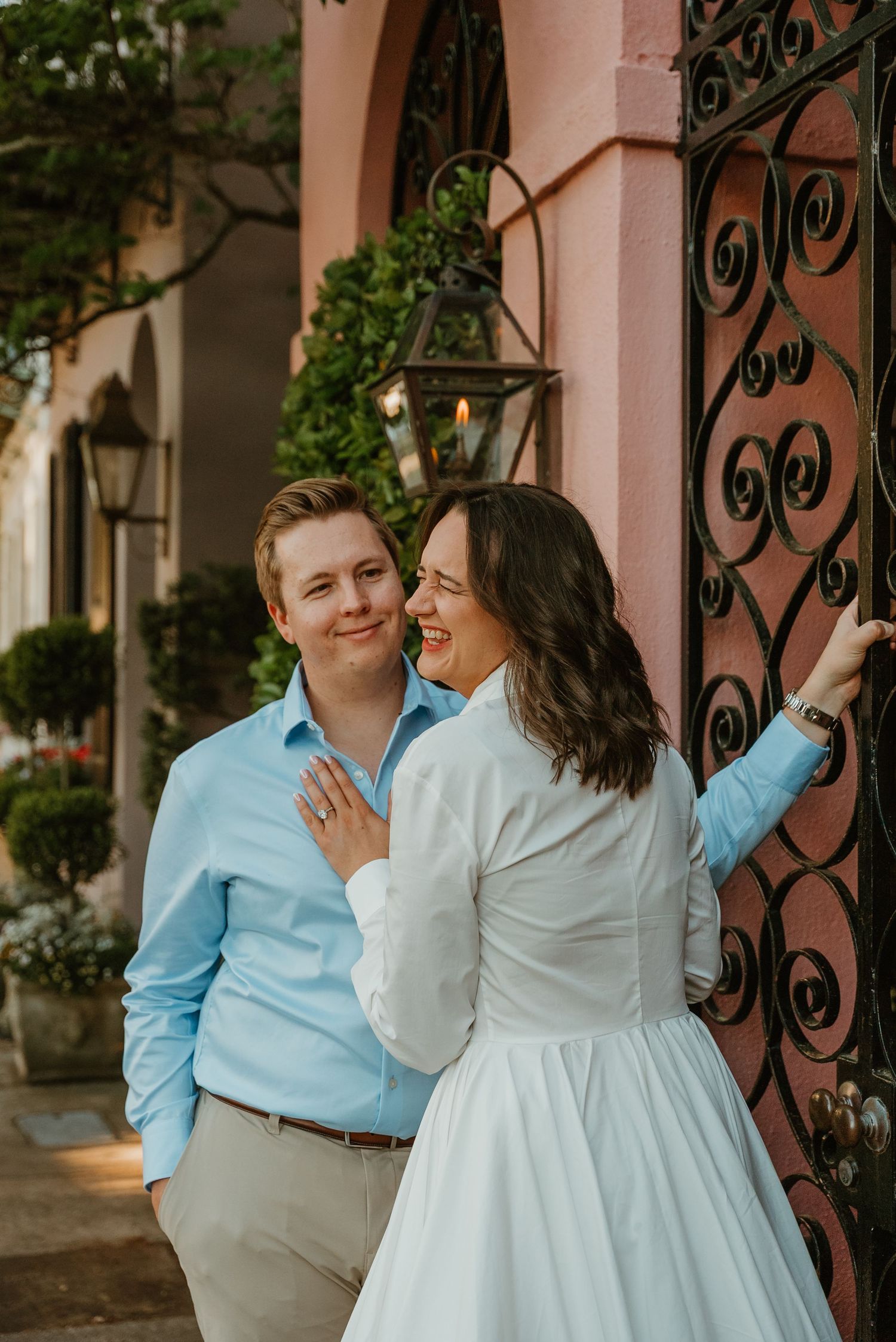 Couple smiling together beside an ornate iron gate at a pink building entrance.