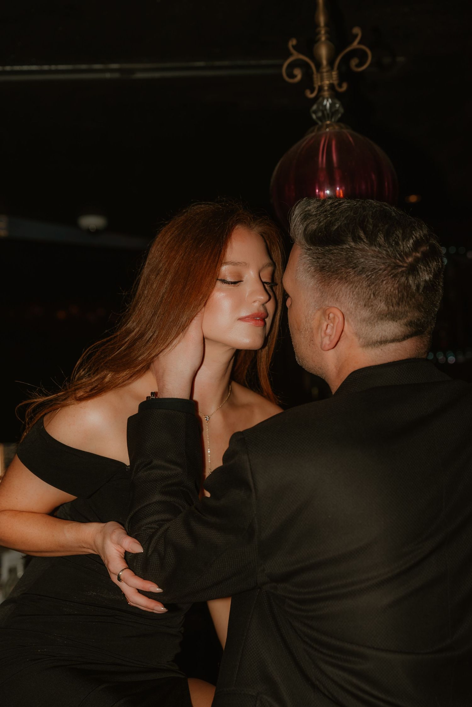 Man in black suit tenderly holding a redhead woman's face in a moody dark venue.