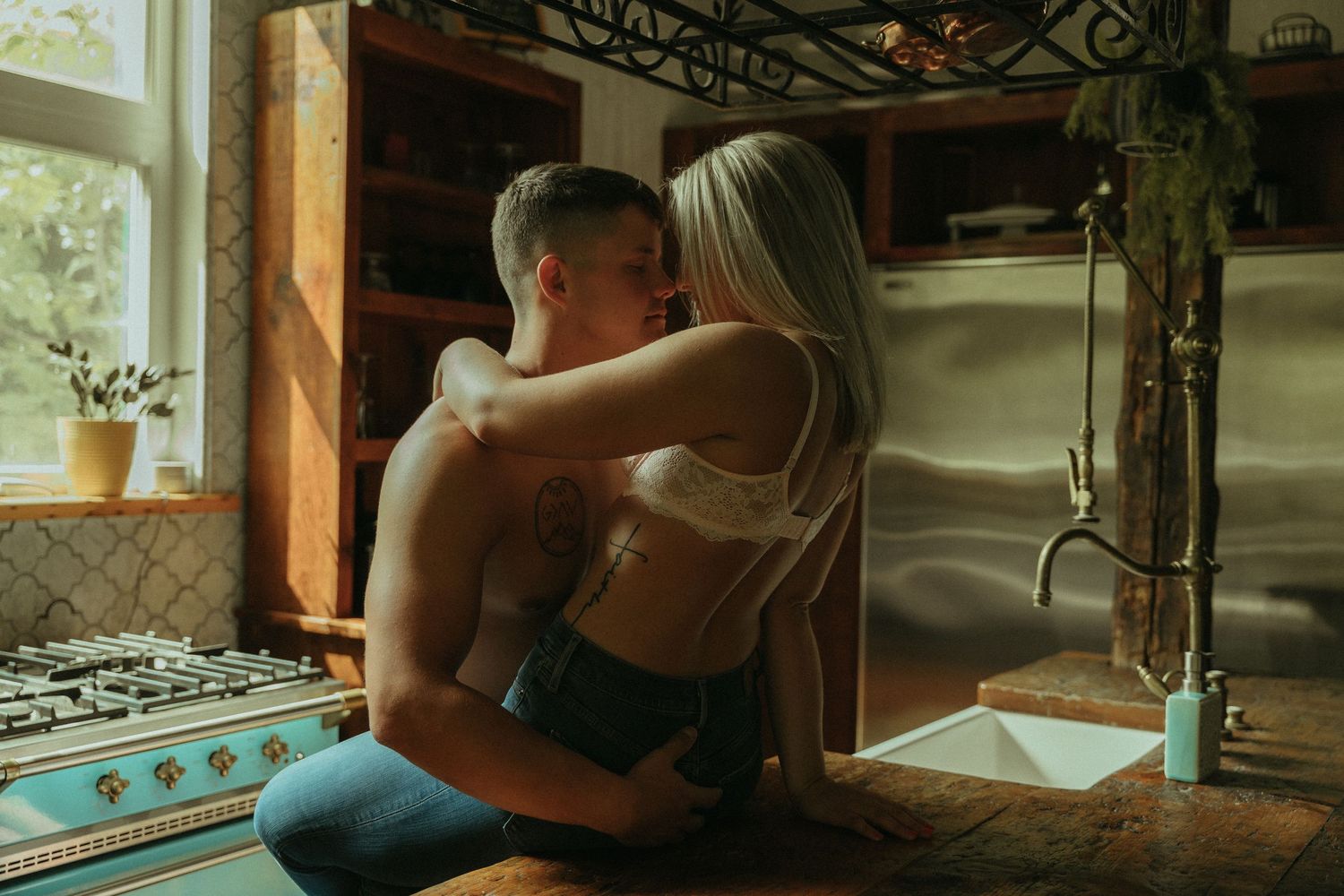 Couple sharing an intimate kiss in a rustic kitchen, man shirtless with tattoos, woman in white lace bralette.