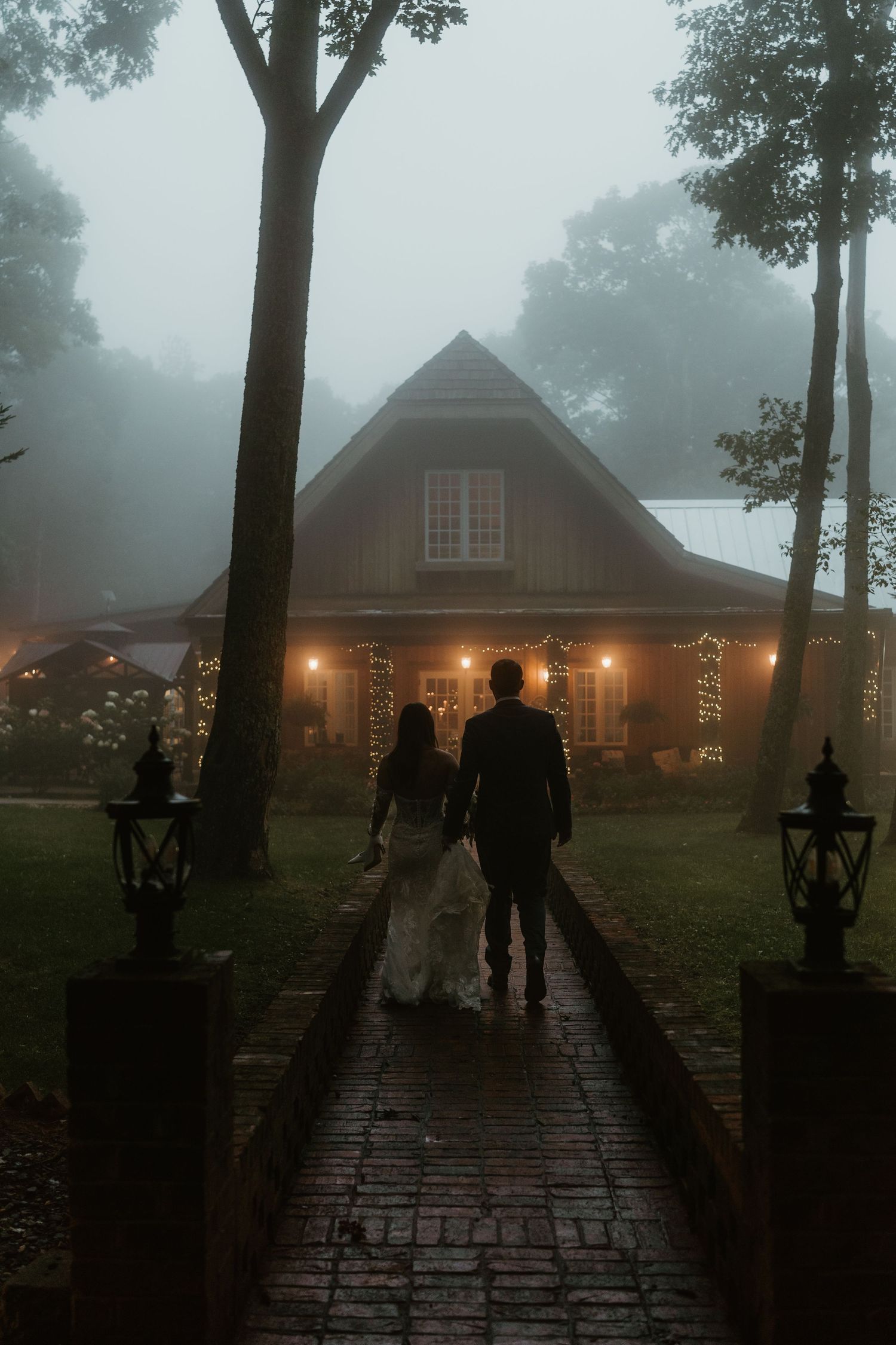 Bride and groom walking hand-in-hand toward warmly lit rustic venue on foggy evening along brick pathway.