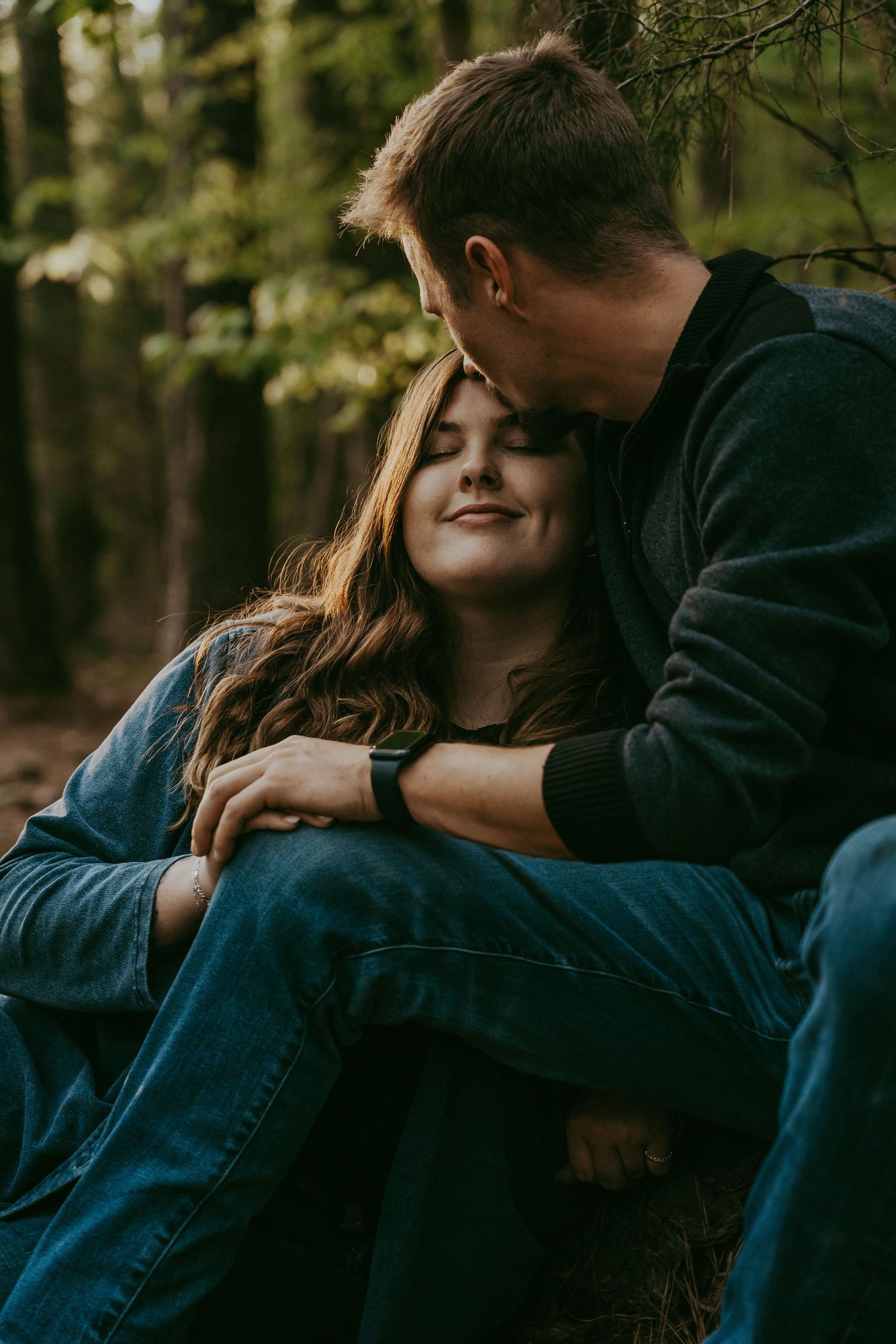 Young couple cuddling tenderly in a forest, woman smiling with eyes closed as man kisses her head.
