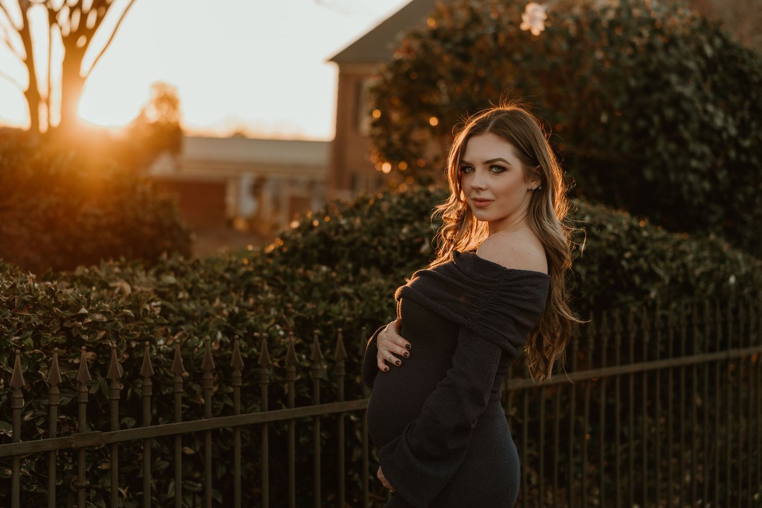 Elegant woman in black off-shoulder dress posing at golden hour sunset near an iron fence and hedges.