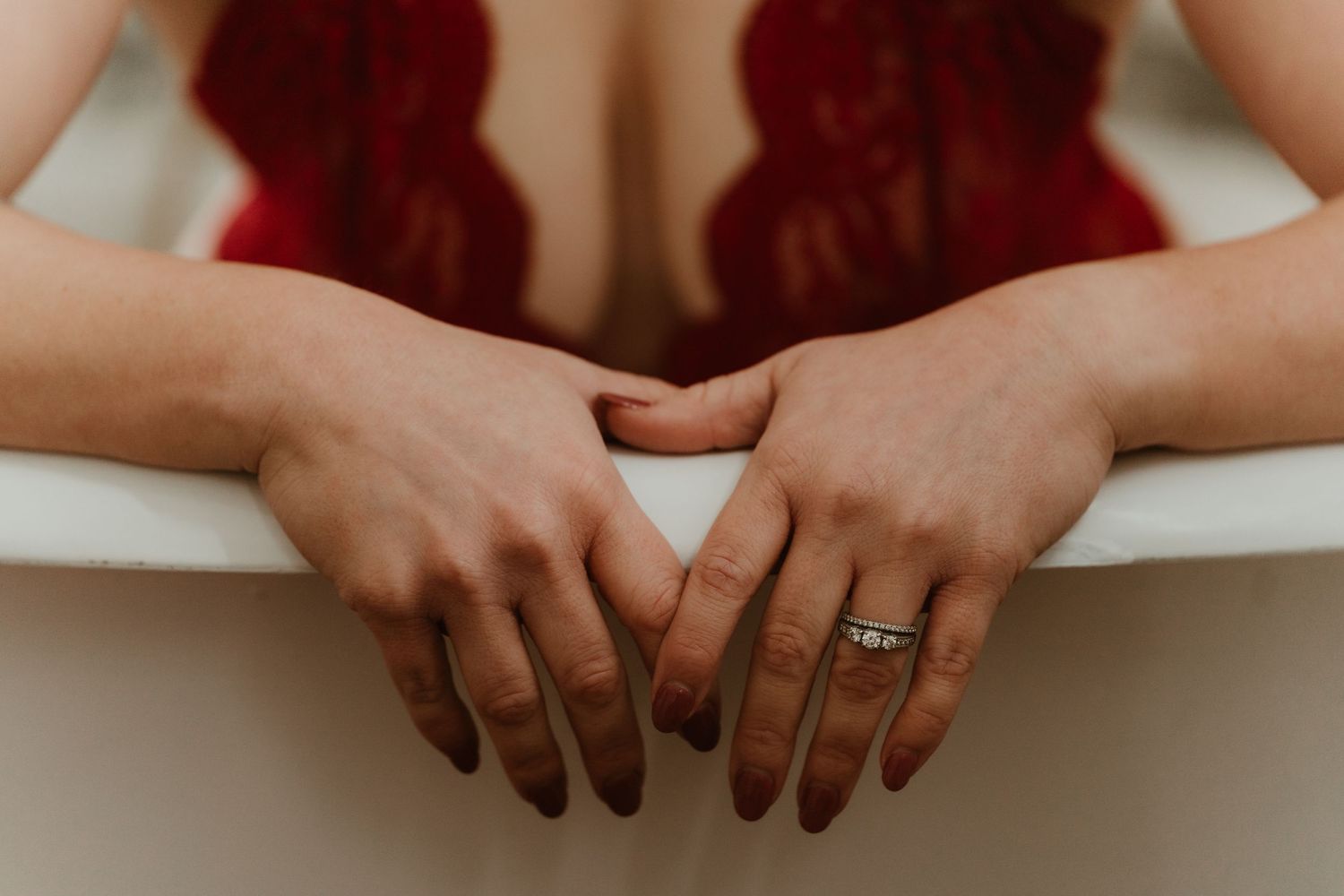 Close-up of a woman's hands resting on a white bathtub edge, wearing a silver ring, with red lingerie reflected.