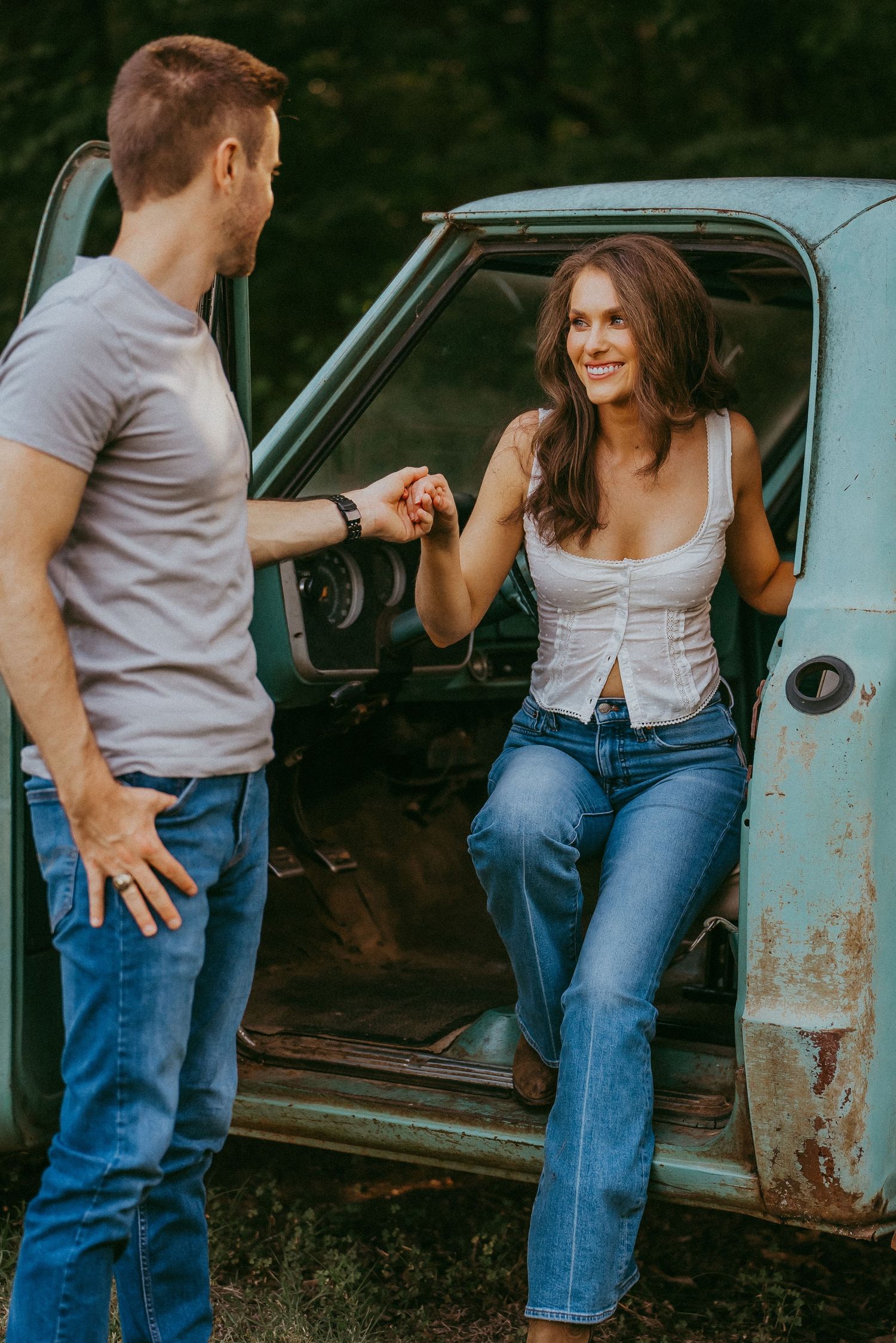 Smiling woman sitting in a vintage rusty truck door while man holds her hand outdoors.
