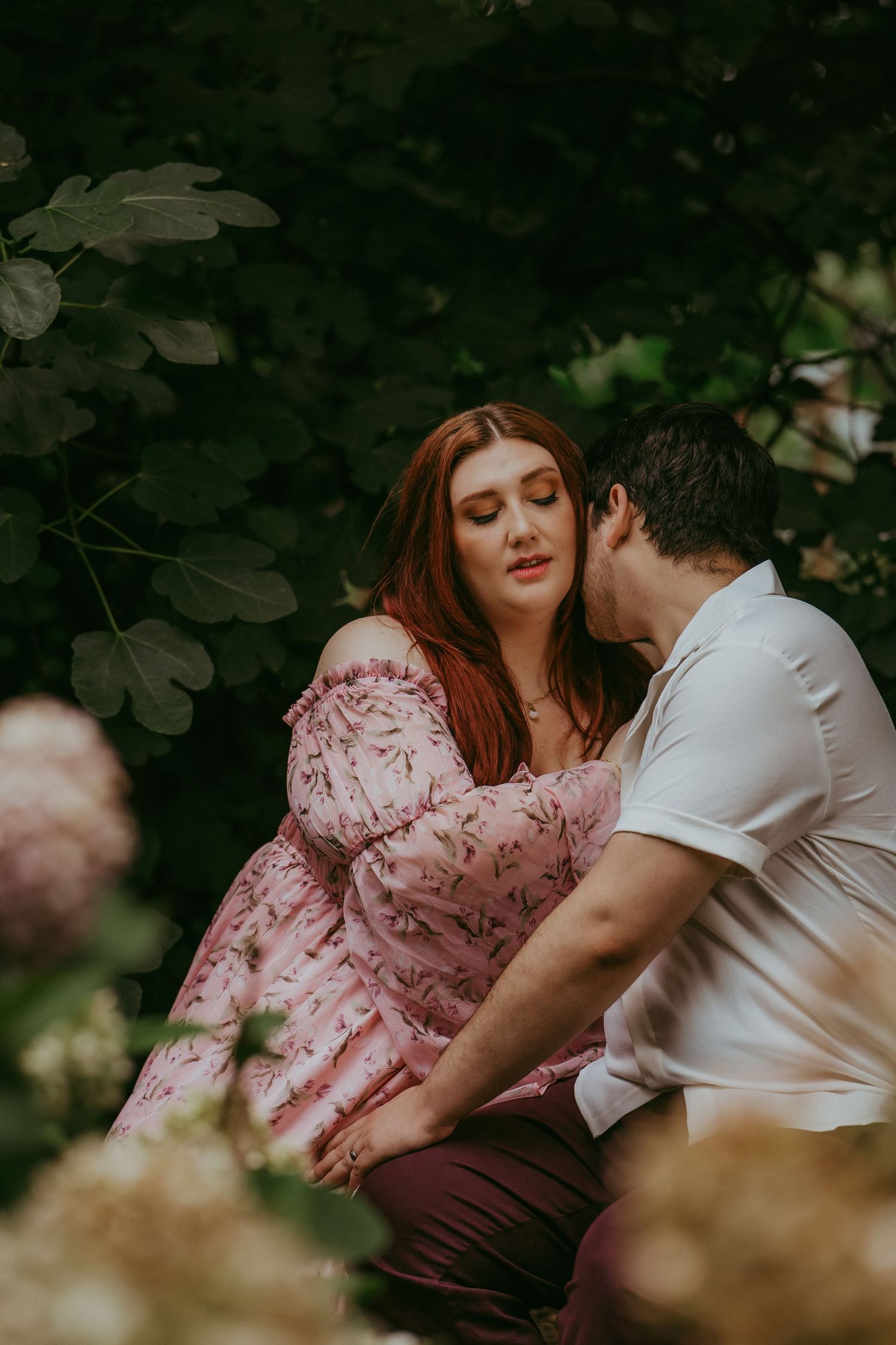 Romantic couple sitting among hydrangeas and fig trees, man kissing womans shoulder in a garden.