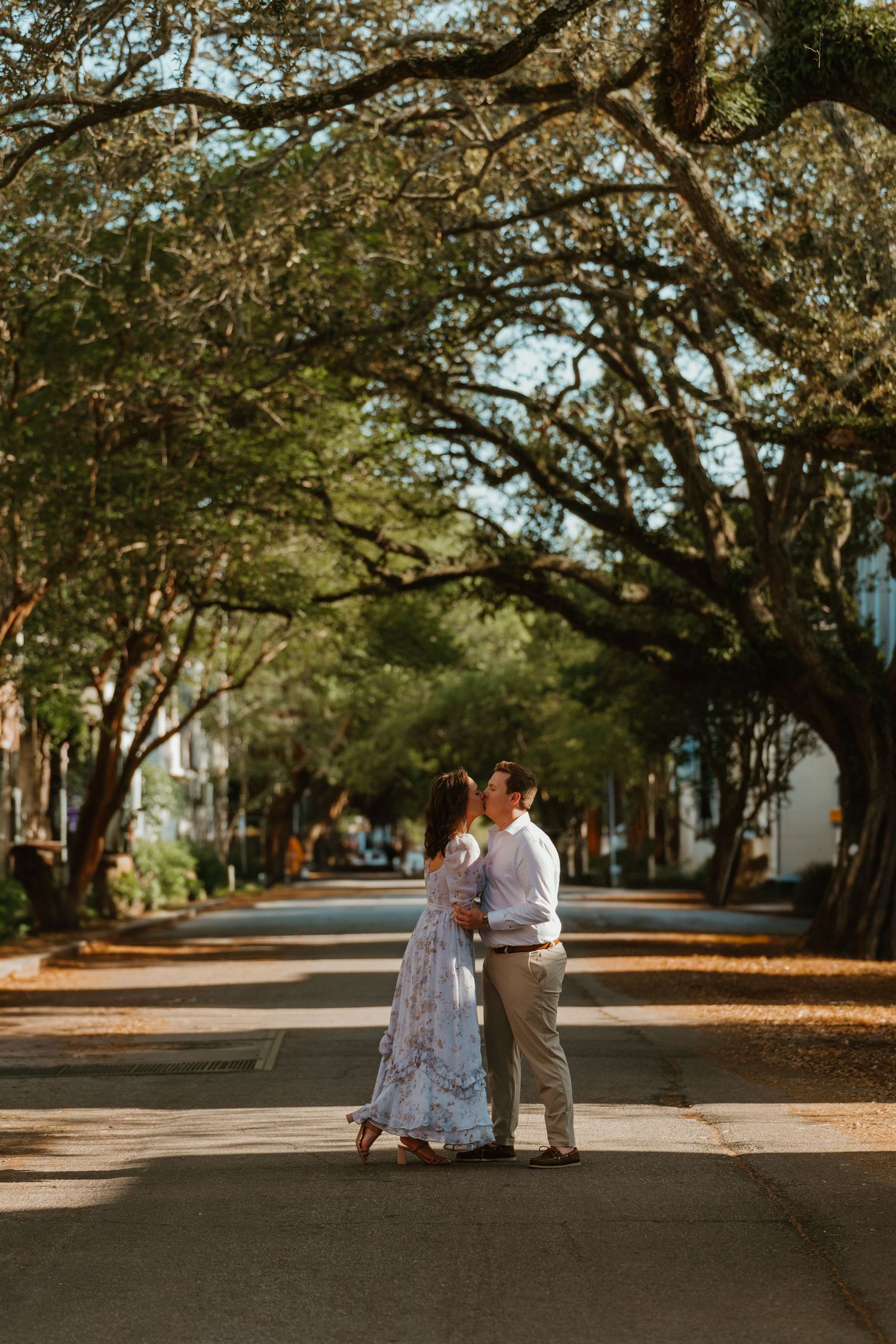 Couple sharing a romantic kiss on a tree-lined street with sprawling oak trees during golden hour engagement session.