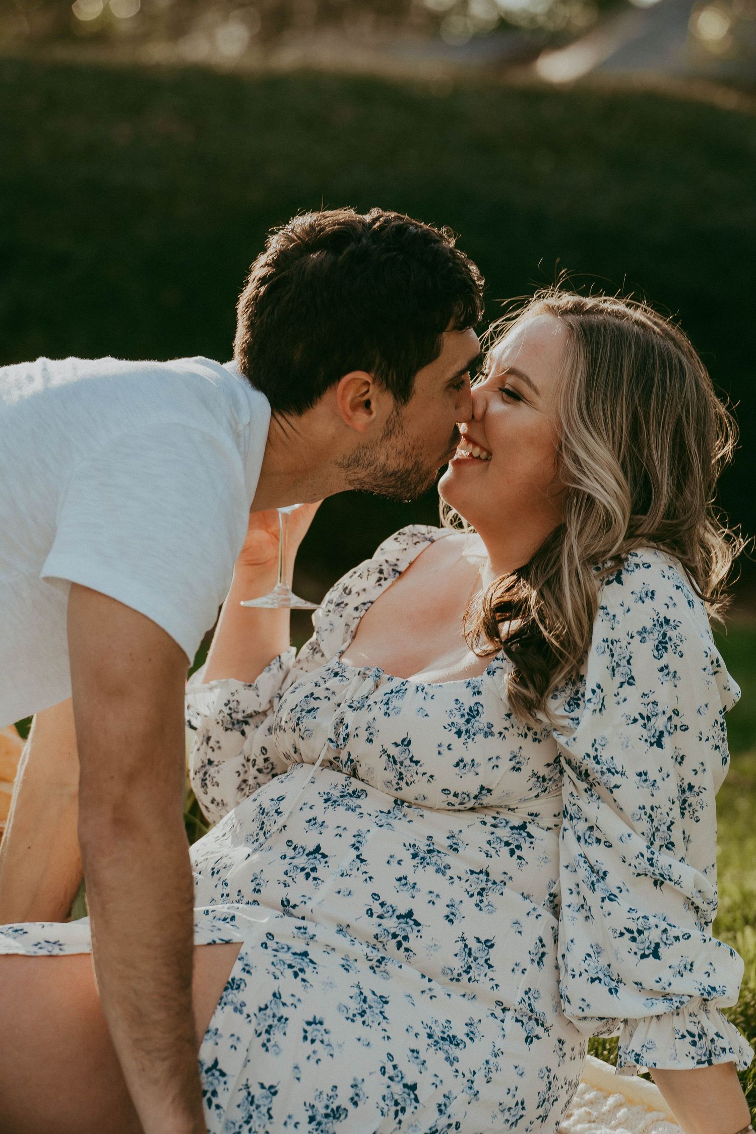 Man kissing smiling woman on cheek outdoors, both wearing casual attire in warm sunlight.