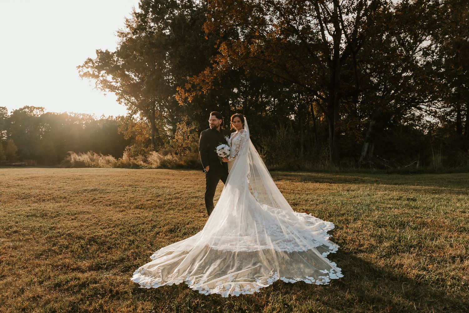 Bride and groom embracing in golden hour sunlight with dramatic lace cathedral veil trailing across grass.