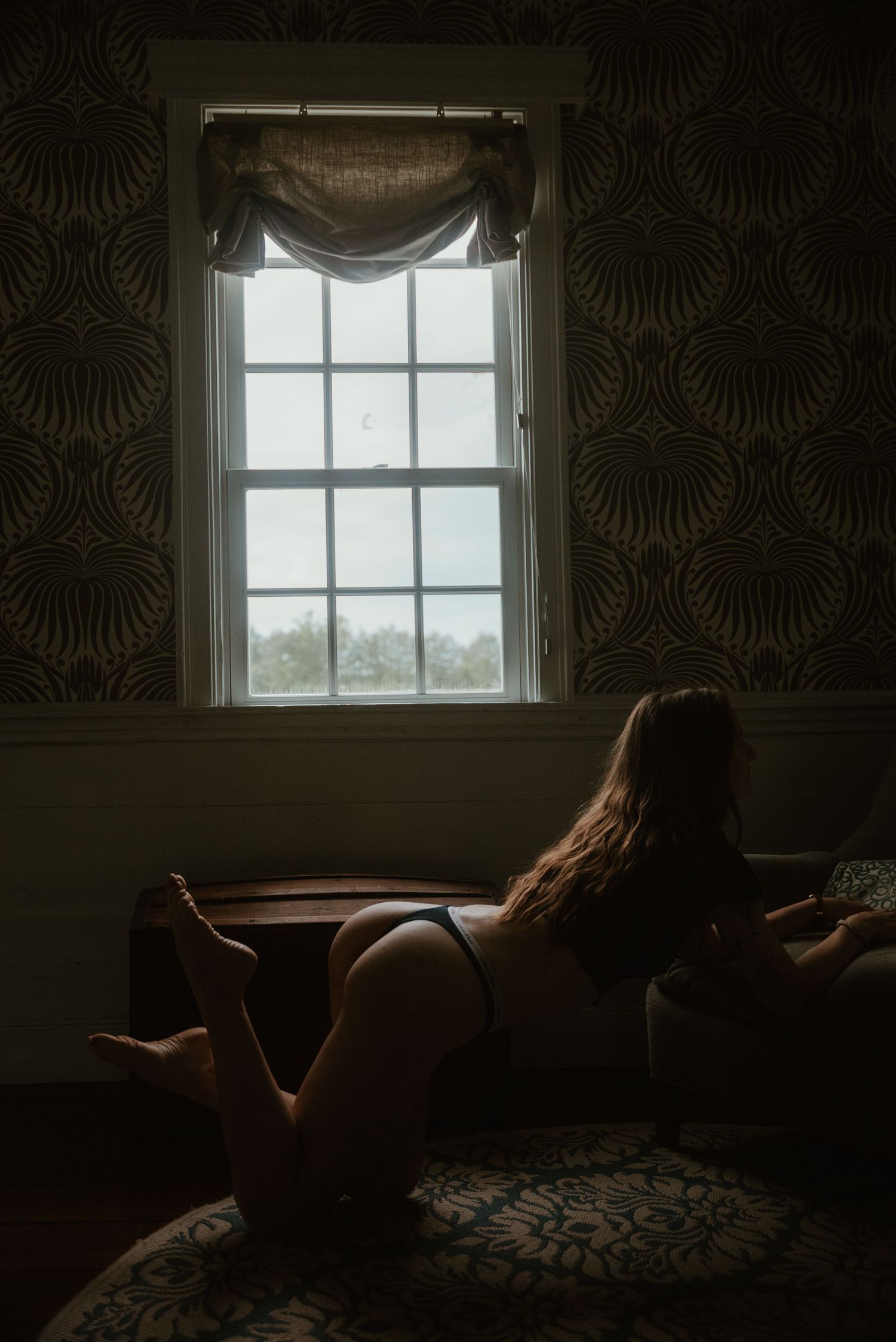 Woman in black lingerie reclining on a chair facing a large patterned wallpaper window.