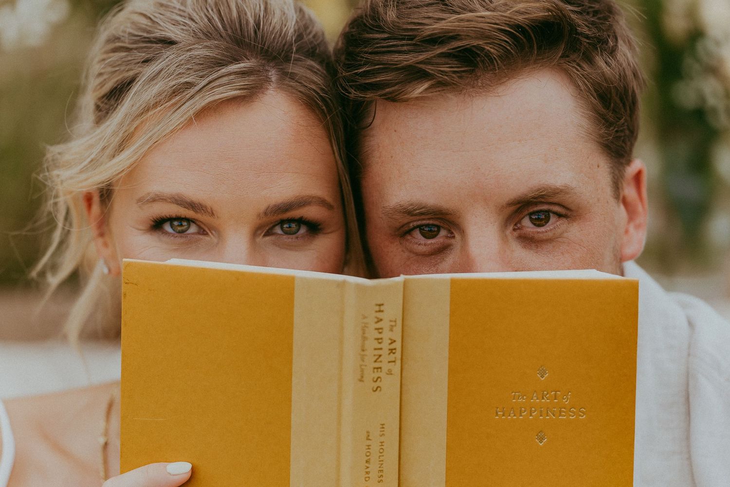 Smiling couple peeking over a yellow vintage book during a romantic outdoor engagement photo session.